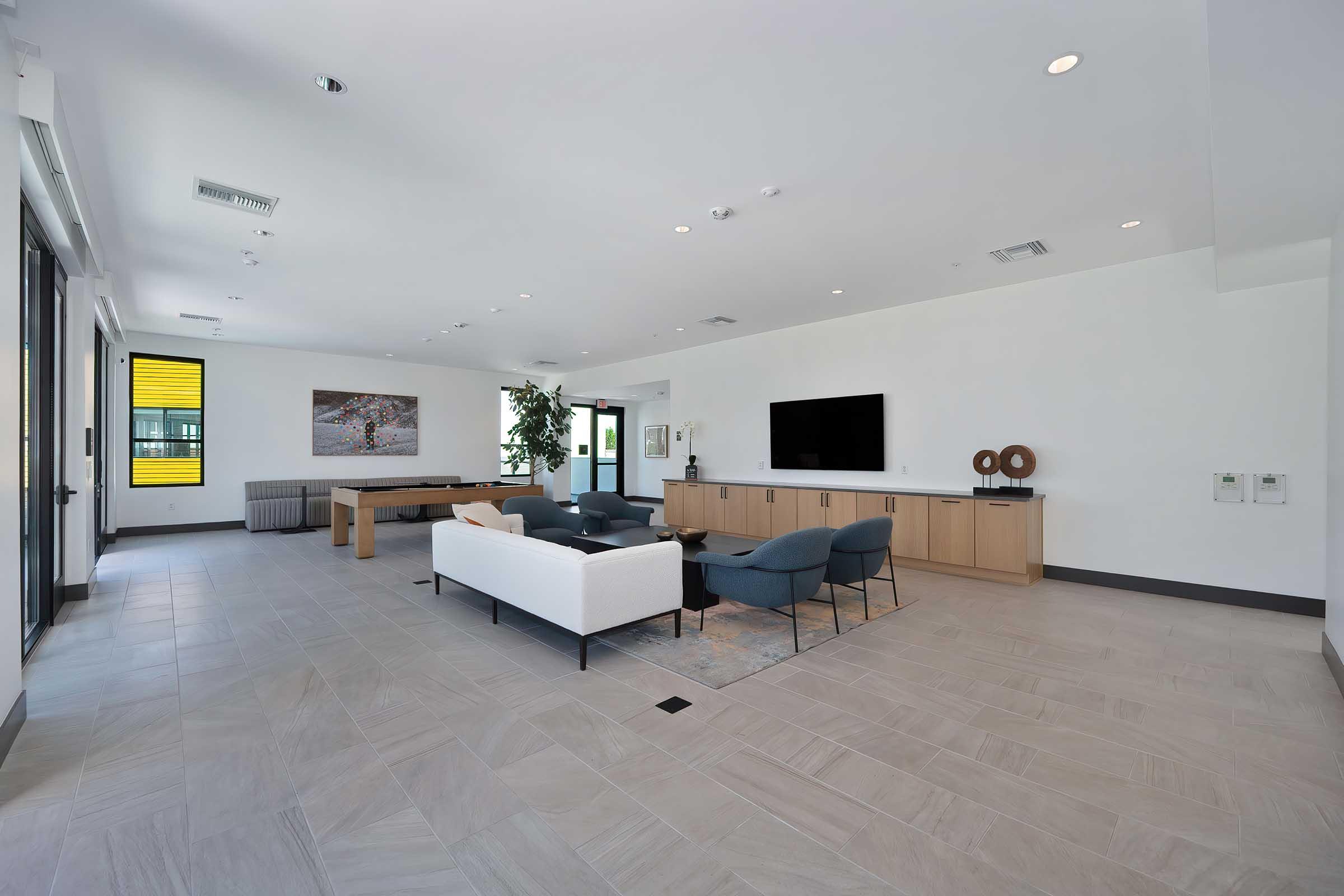 A modern, spacious lounge area featuring a light-colored sofa, blue chairs, and a wooden dining table. There’s a large flat-screen TV on a minimalist cabinet, large windows allowing natural light, and a potted plant for a touch of greenery. The flooring is tiled with a light, neutral color.