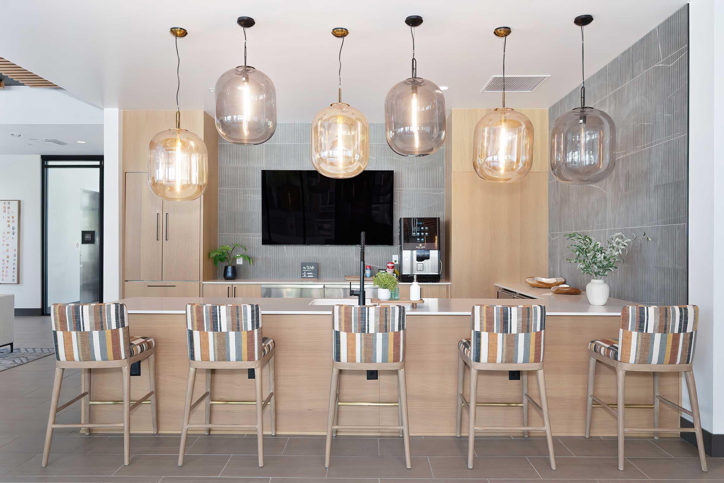 Modern kitchen interior featuring a sleek wooden island with high stools adorned in striped fabric. Above the island hang several stylish glass pendant lights. A large flat-screen TV is mounted on the wall, and plants and decorative items add a touch of elegance to the space.