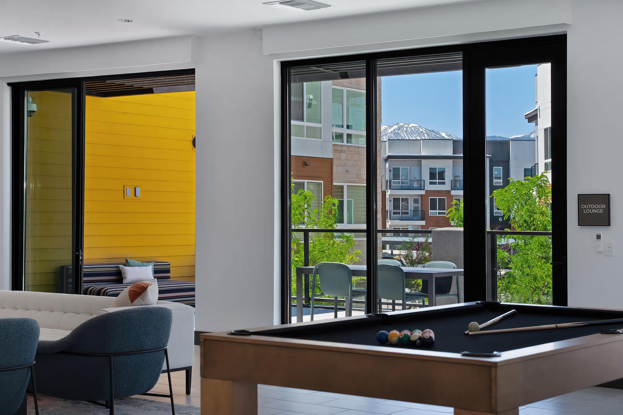 Interior view of a modern lounge area featuring a pool table, large windows with natural light, and a balcony with outdoor seating. The background shows a colorful building facade and green trees, with distant mountains visible through the window.