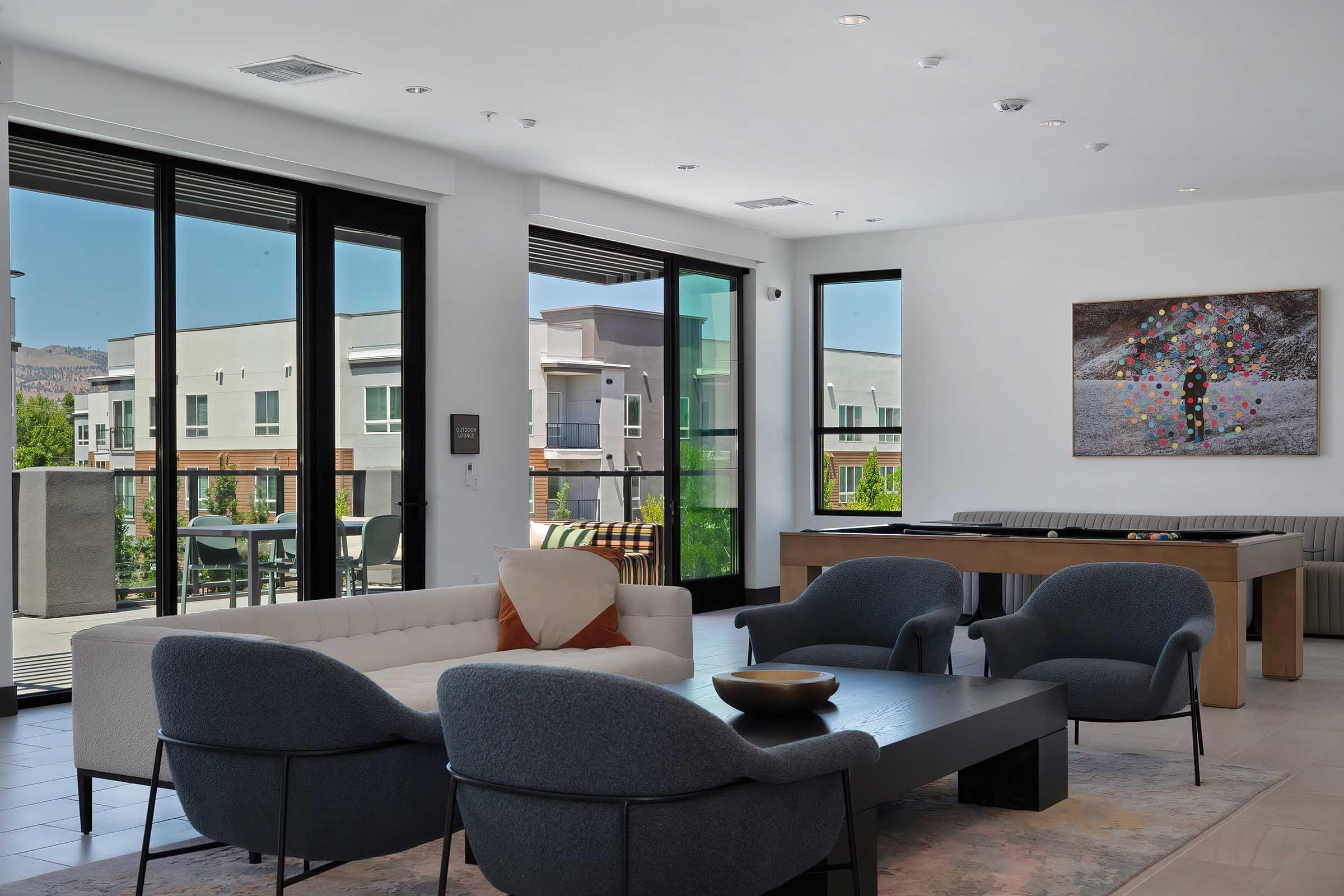 A modern living space featuring large windows with views of buildings outside. The room has a comfortable sofa, two stylish armchairs, and a sleek coffee table. A colorful artwork is hanging on the wall, and there's a dining table off to the side. The decor is contemporary with light colors and minimalistic design.