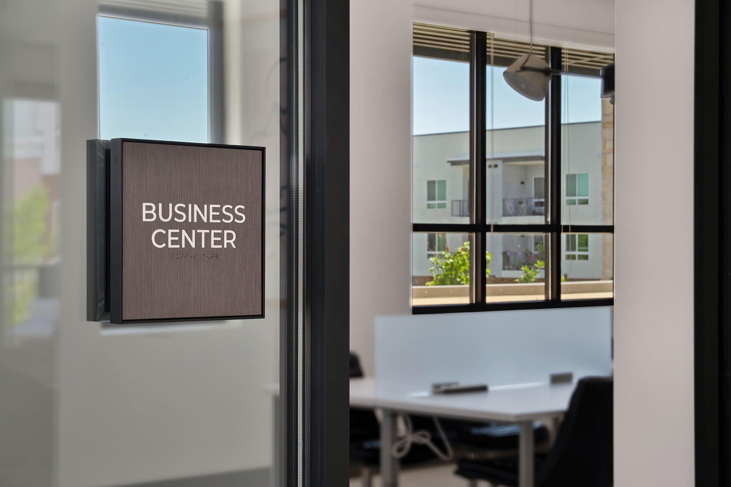 Sign reading "BUSINESS CENTER" on a modern office door, with large windows showcasing a view of an outdoor area. Inside the office space, there are desks and black office chairs, creating a professional environment.