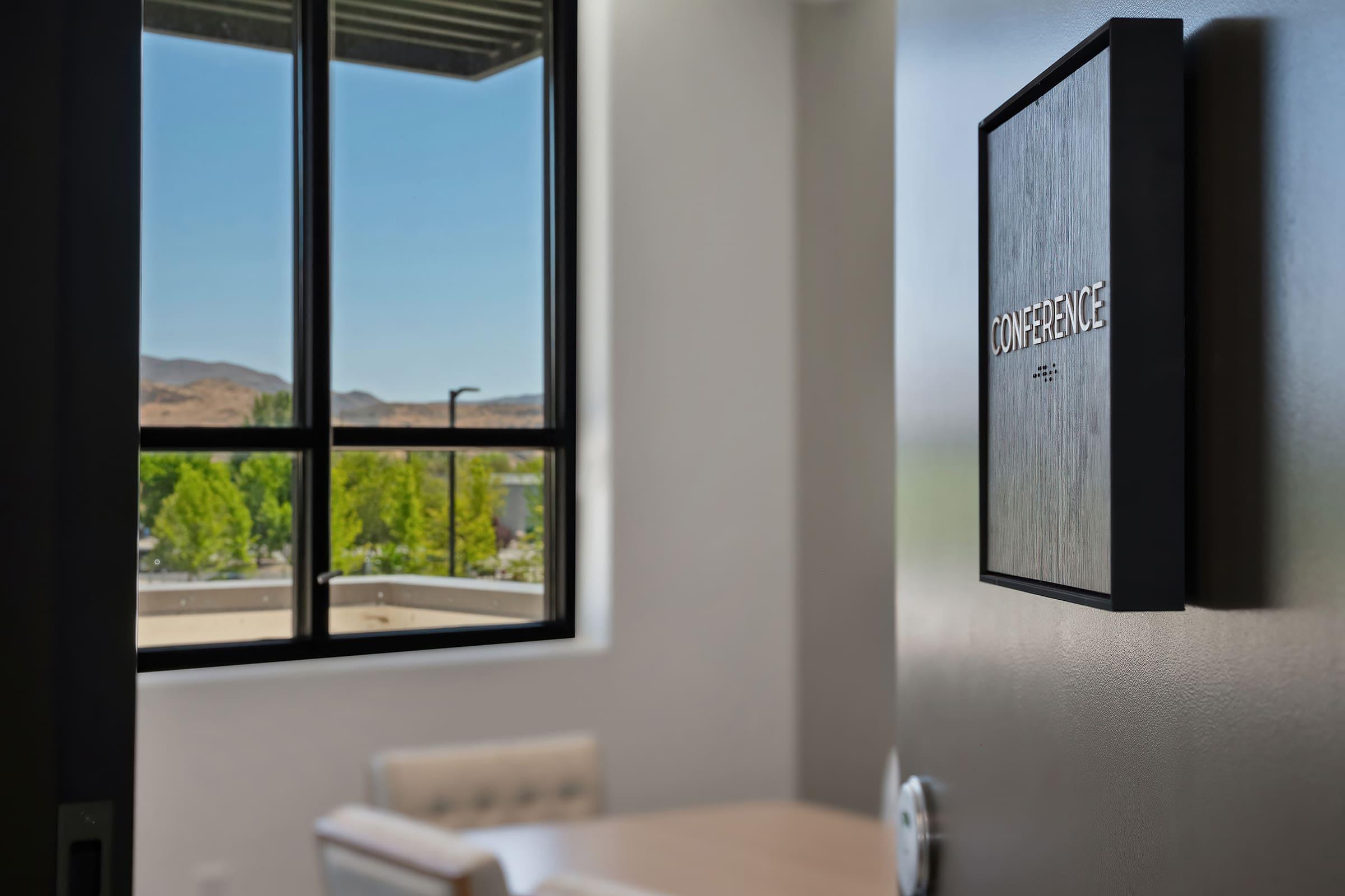 A close-up view of a door to a conference room, featuring a sign that reads "CONFERENCE." In the background, a large window shows a clear blue sky and green trees, with a glimpse of mountains beyond. The focus is on the door sign, while the interior of the room is softly blurred.