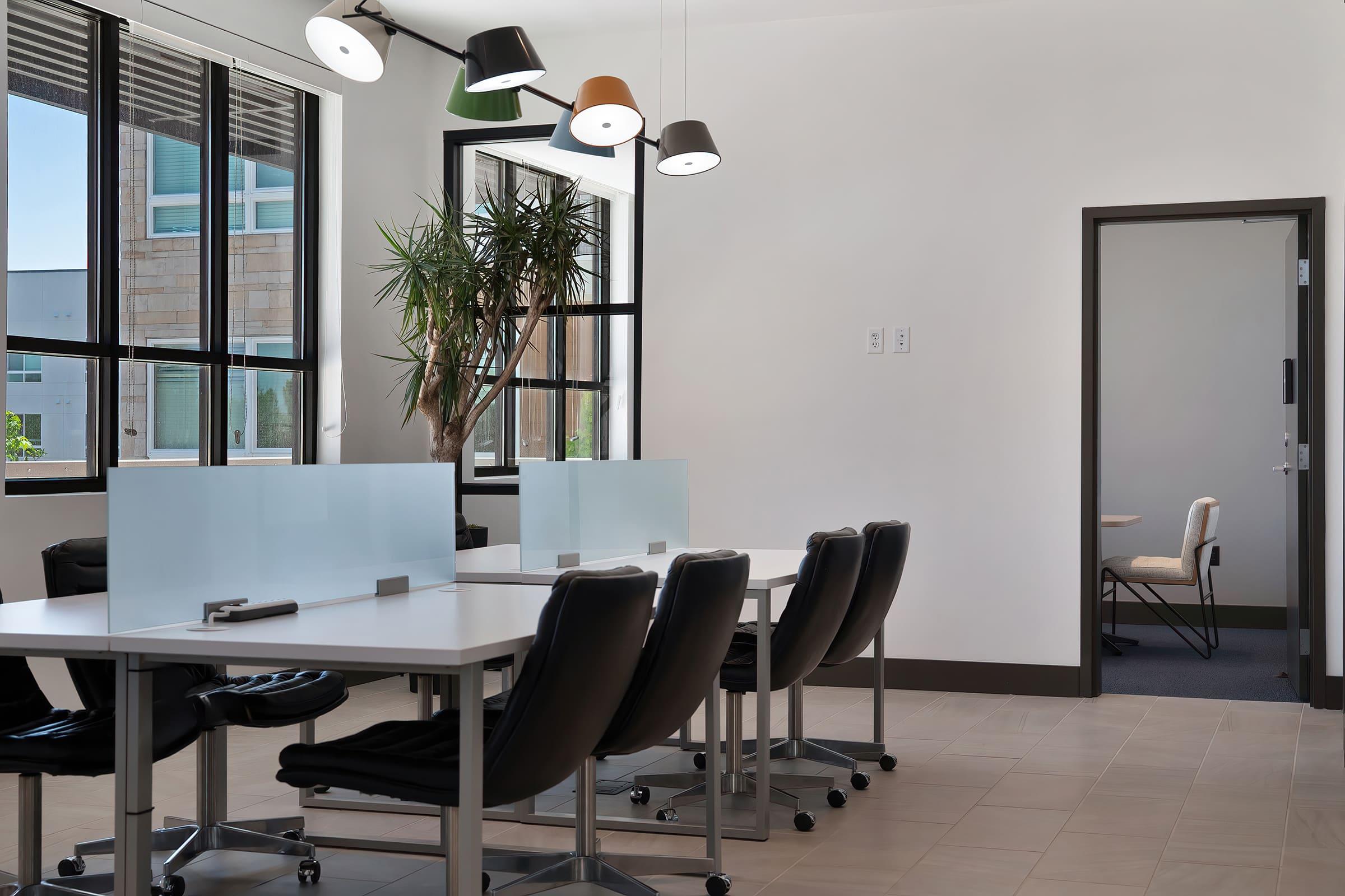 Modern office interior featuring multiple white desks with black ergonomic chairs. There are glass partitions for privacy, and large windows allowing natural light. Pendant lamps hang from the ceiling, and a potted plant adds a touch of greenery. A door leads to another room, visible in the background.