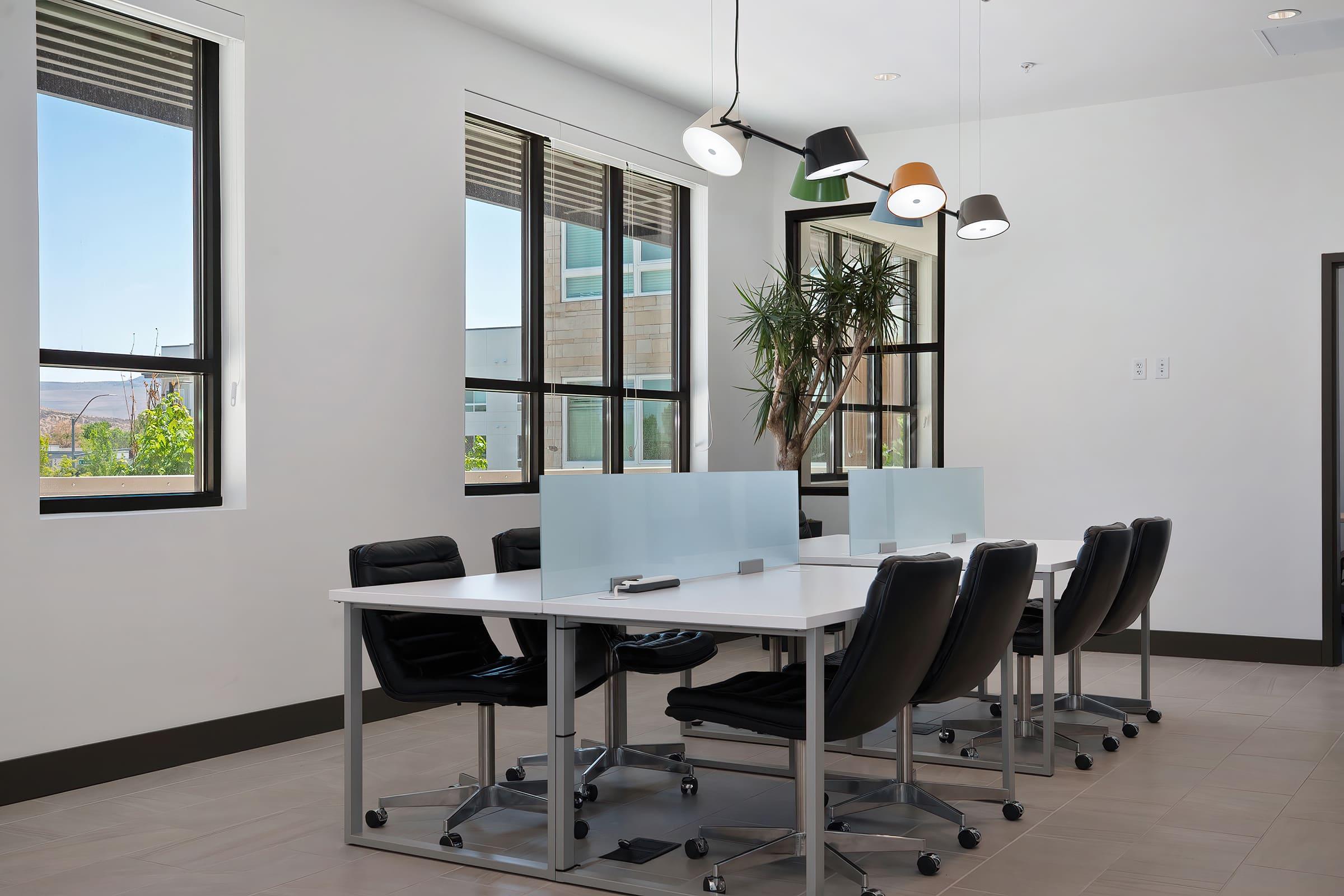 A bright and modern office space featuring a large meeting table with black rolling chairs. The room has large windows allowing natural light in, and unique pendant lights hang from the ceiling. There are green plants in the room, creating a fresh and inviting atmosphere.
