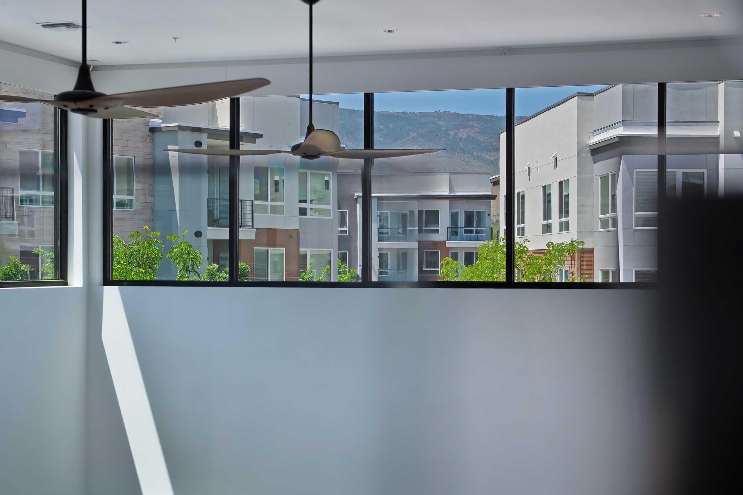 Interior view of a modern apartment with large windows showcasing neighboring buildings and a mountain backdrop. The space features minimalistic design elements, including ceiling fans and a bright atmosphere. Sunlight streams in, illuminating the clean lines of the walls and architecture.