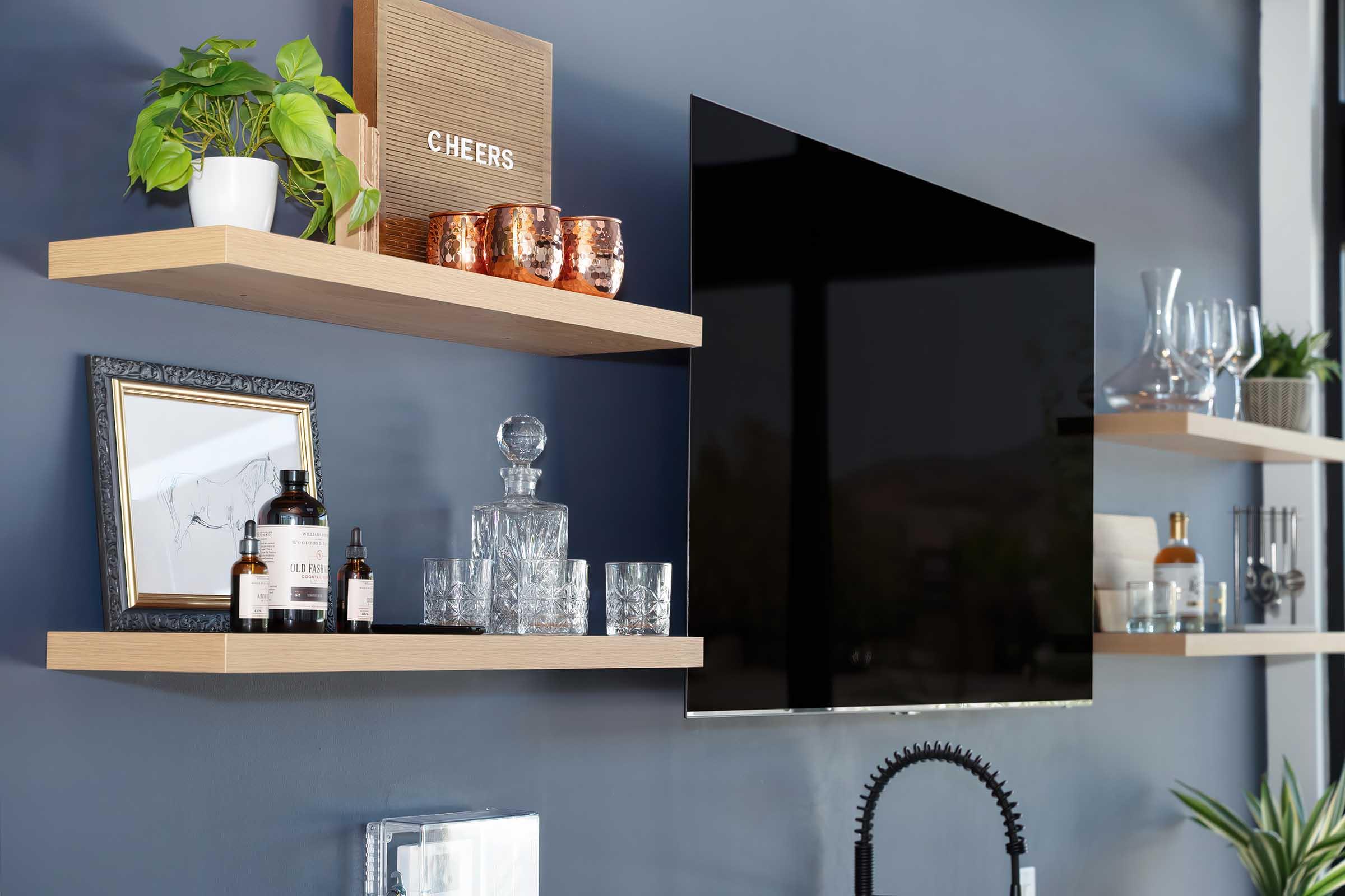 A modern living space with a flat-screen TV mounted on a dark wall. Above it, there are wooden shelves displaying various decorative items, including a plant, copper mugs, a framed picture, a glass decanter, and multiple glassware. A stylish "CHEERS" sign adds a touch of personality.