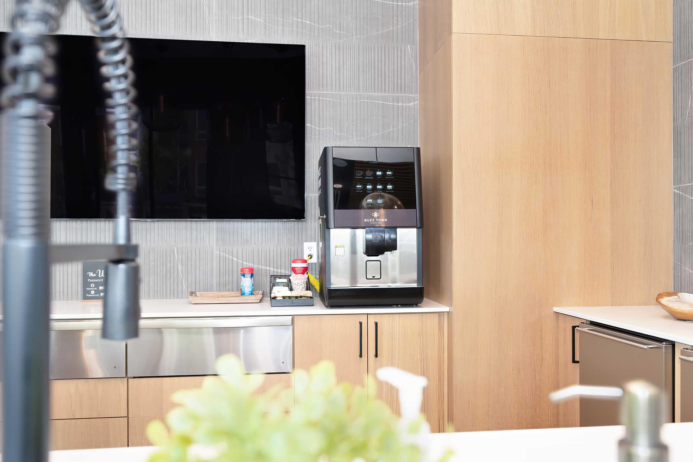 A modern kitchen setting featuring a coffee machine on a countertop, with a TV mounted on the wall above. The cabinetry is light wood, and a small succulent plant is visible in the foreground, adding a touch of greenery to the space. The overall aesthetic is sleek and contemporary.