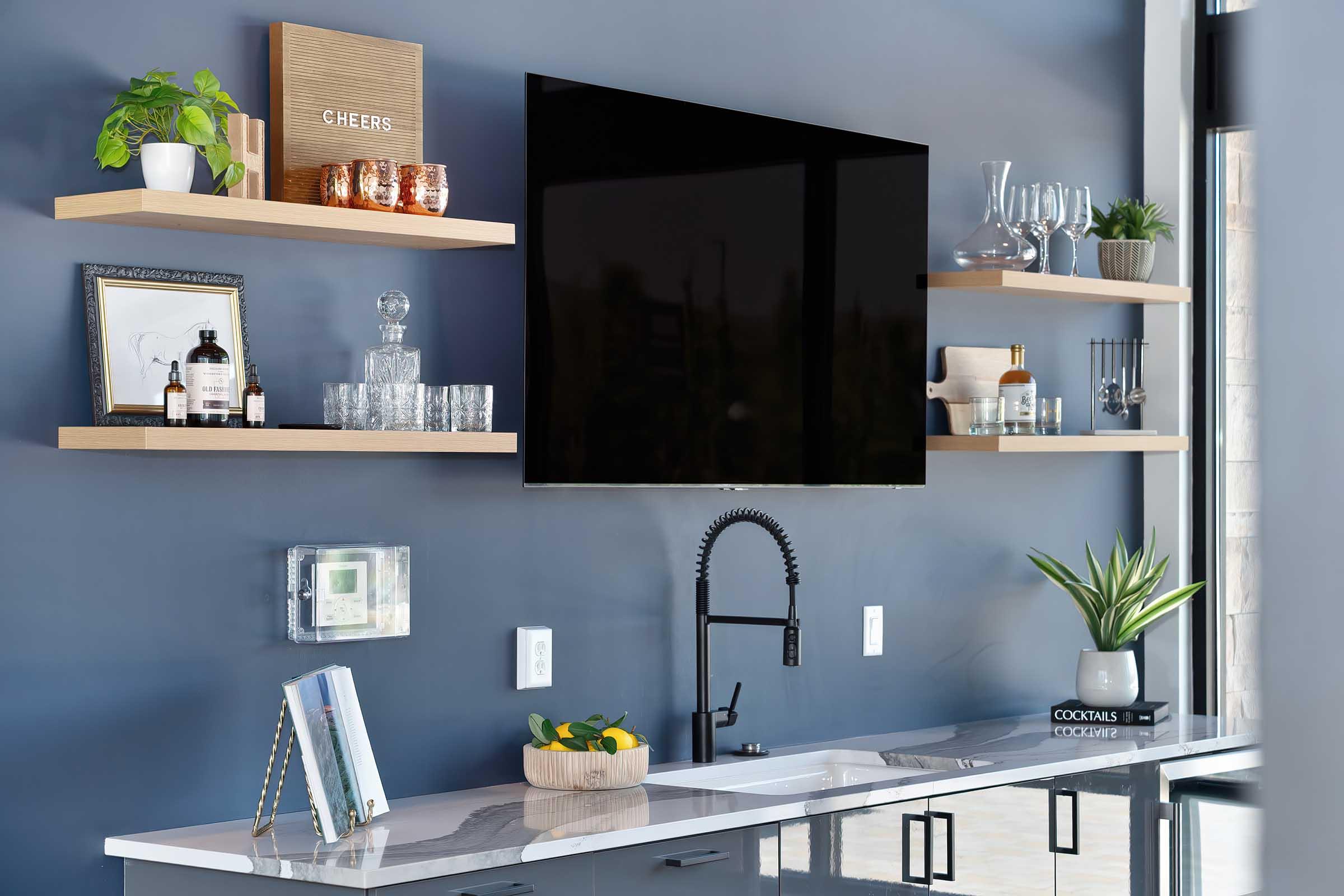 Modern kitchen scene featuring a sleek black television mounted on a blue wall. Floating wooden shelves display a "CHEERS" sign, glassware, plants, and decorative items. A stylish sink with a matte black faucet is positioned on a marble countertop, with a fruit bowl and various books nearby.