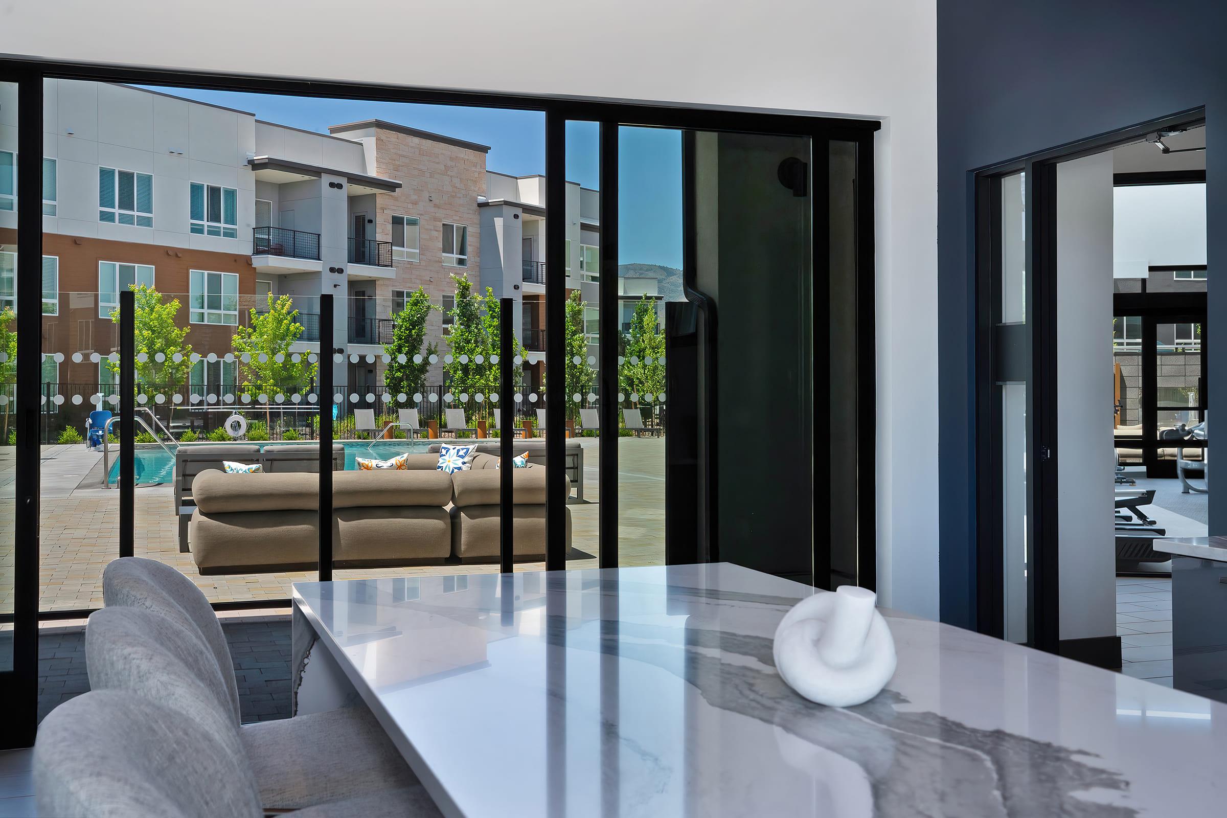 A modern dining area with a marble table and gray chairs, offering a view through large glass doors to a landscaped outdoor patio with seating and greenery. The scene features contemporary architecture in the background, with bright sunlight illuminating the space.