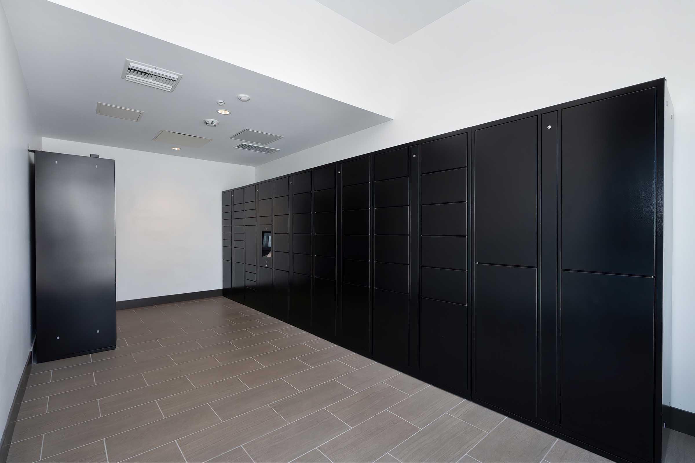 A modern interior space featuring a line of black storage lockers against a light-colored wall, with a tiled floor. The area appears well-lit and minimalistic, suggesting it could be used for package deliveries or personal storage.