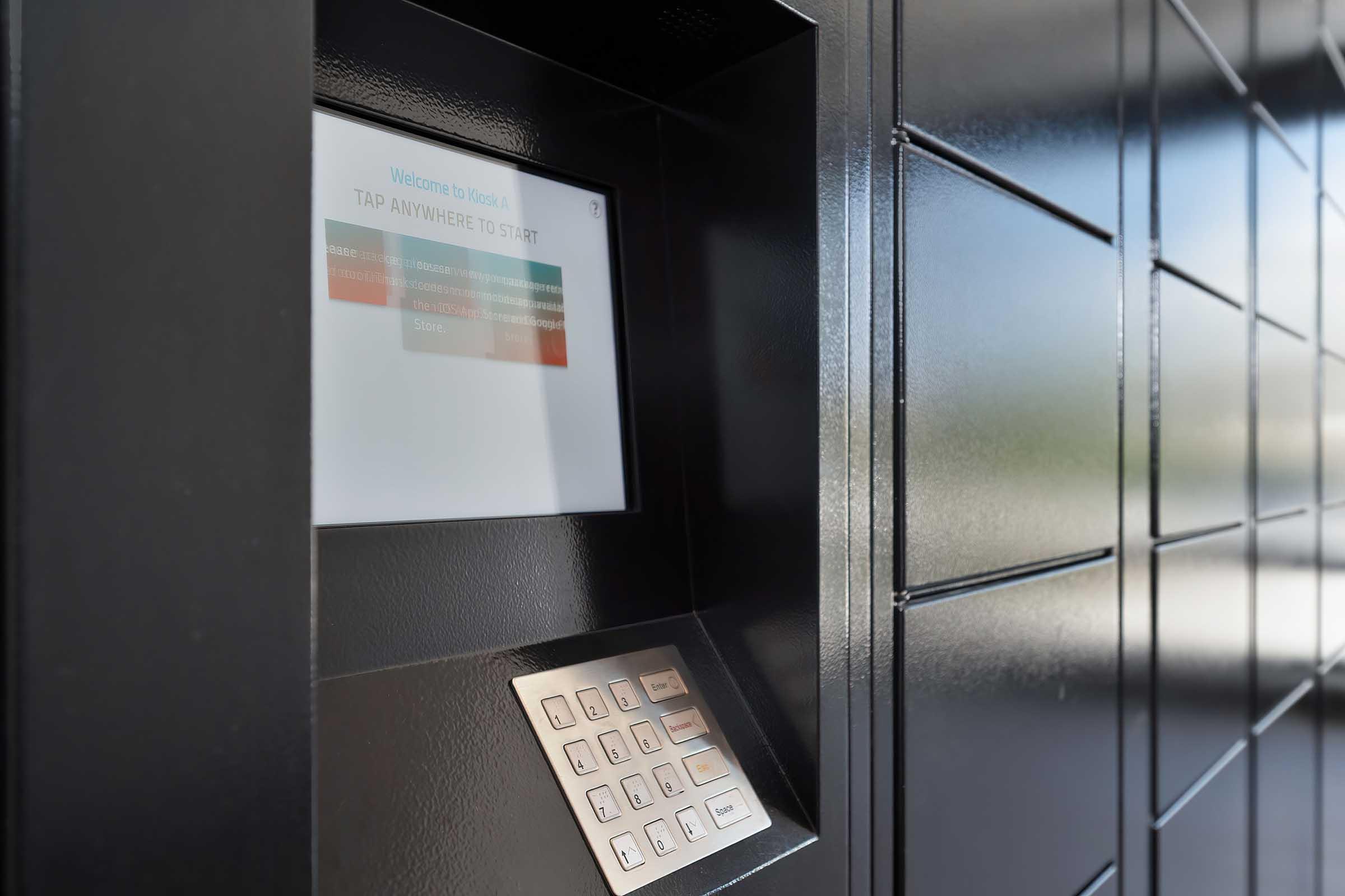 A close-up view of a digital delivery locker interface. The screen displays a welcome message with a prompt to tap anywhere to start, while a keypad with numerical and functional buttons is visible below the screen. The surrounding surface features a sleek, modern design in dark colors.