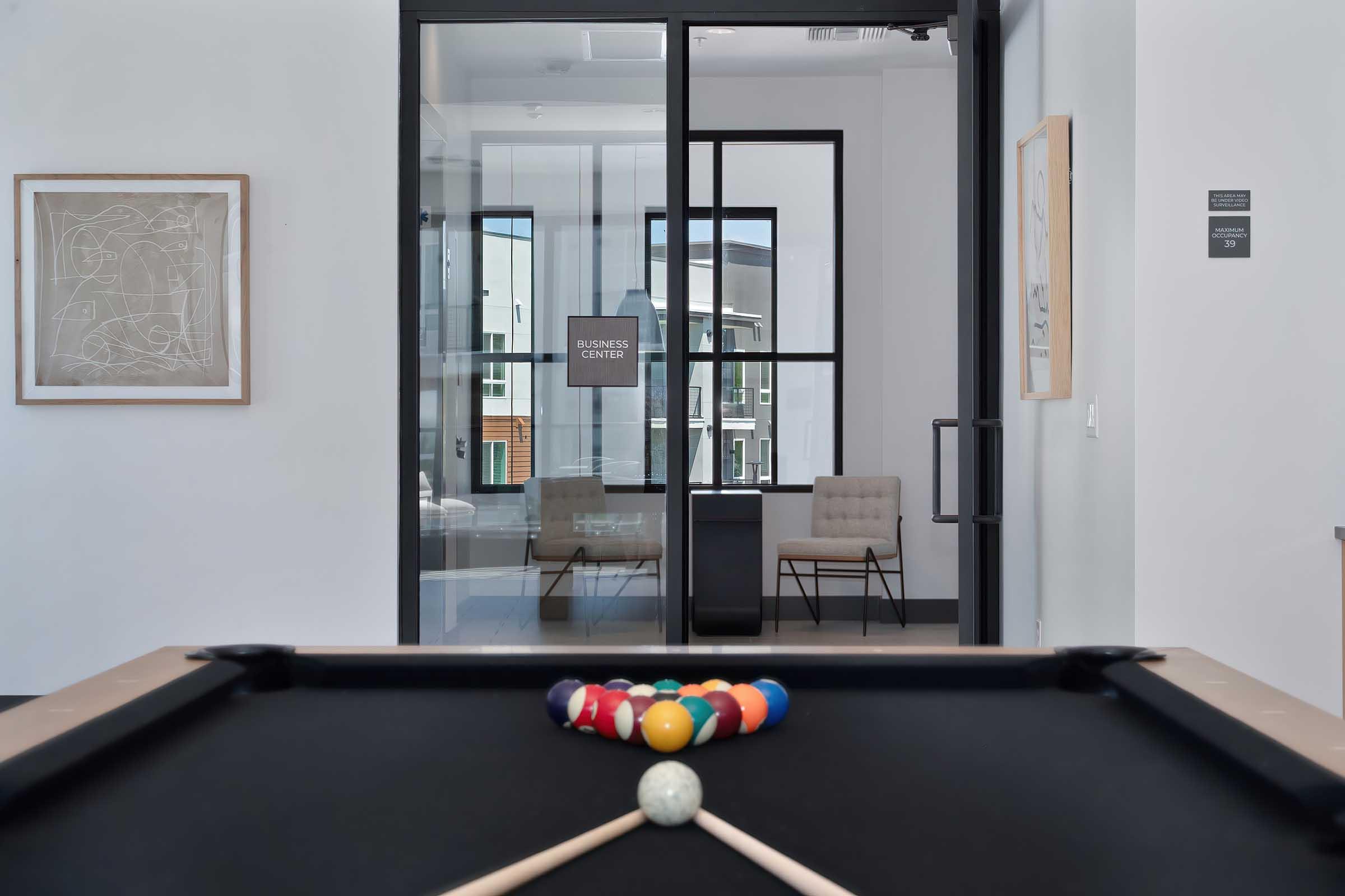A pool table with a cue stick positioned over a set of colorful billiard balls. In the background, a modern business center is visible through large glass doors, featuring minimalist seating and decor in a well-lit, contemporary space.