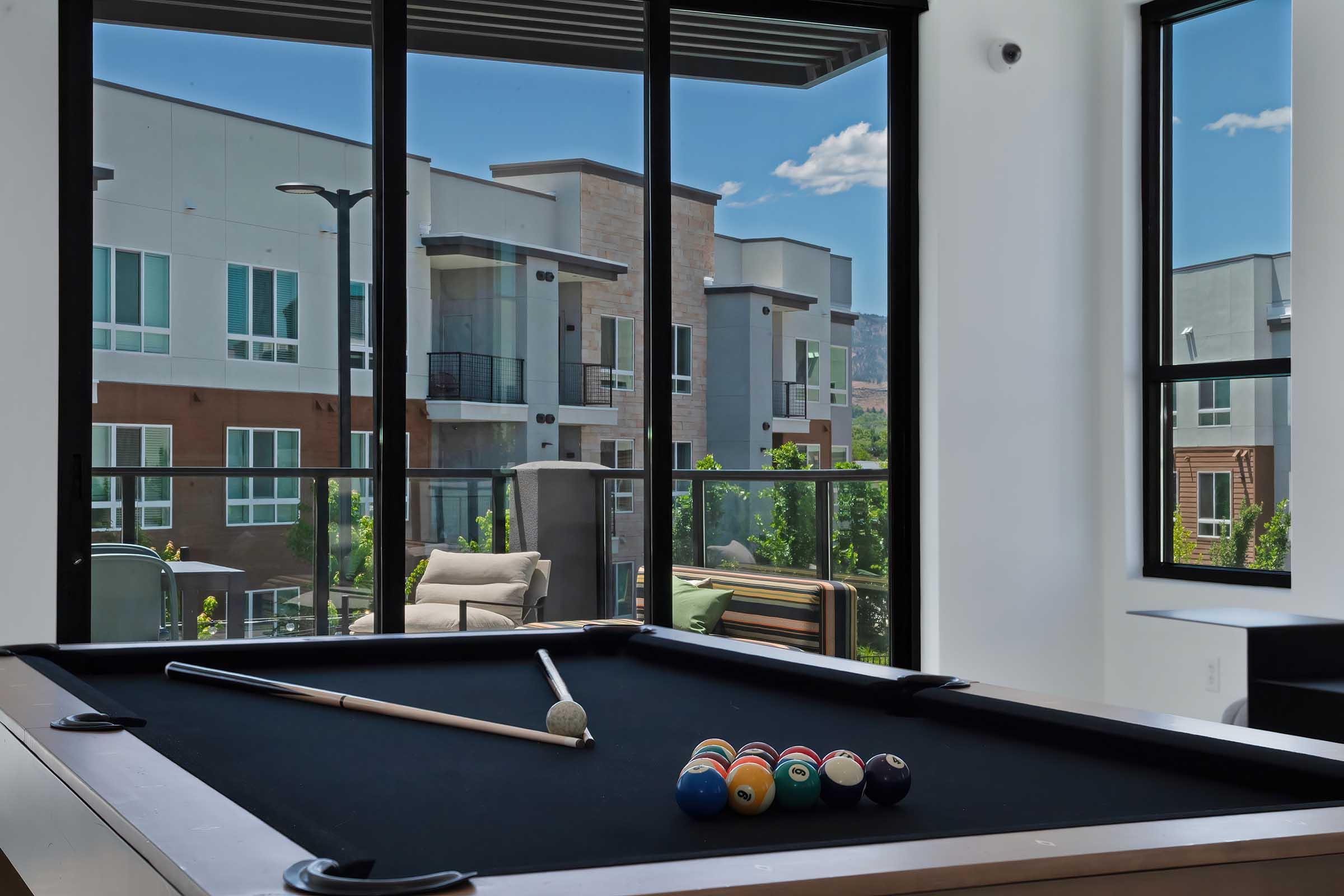A view from a modern room featuring a pool table with a black felt surface, a cue stick, and a set of colored billiard balls. Through large windows, there is a scenic landscape of contemporary apartment buildings and greenery under a clear blue sky.