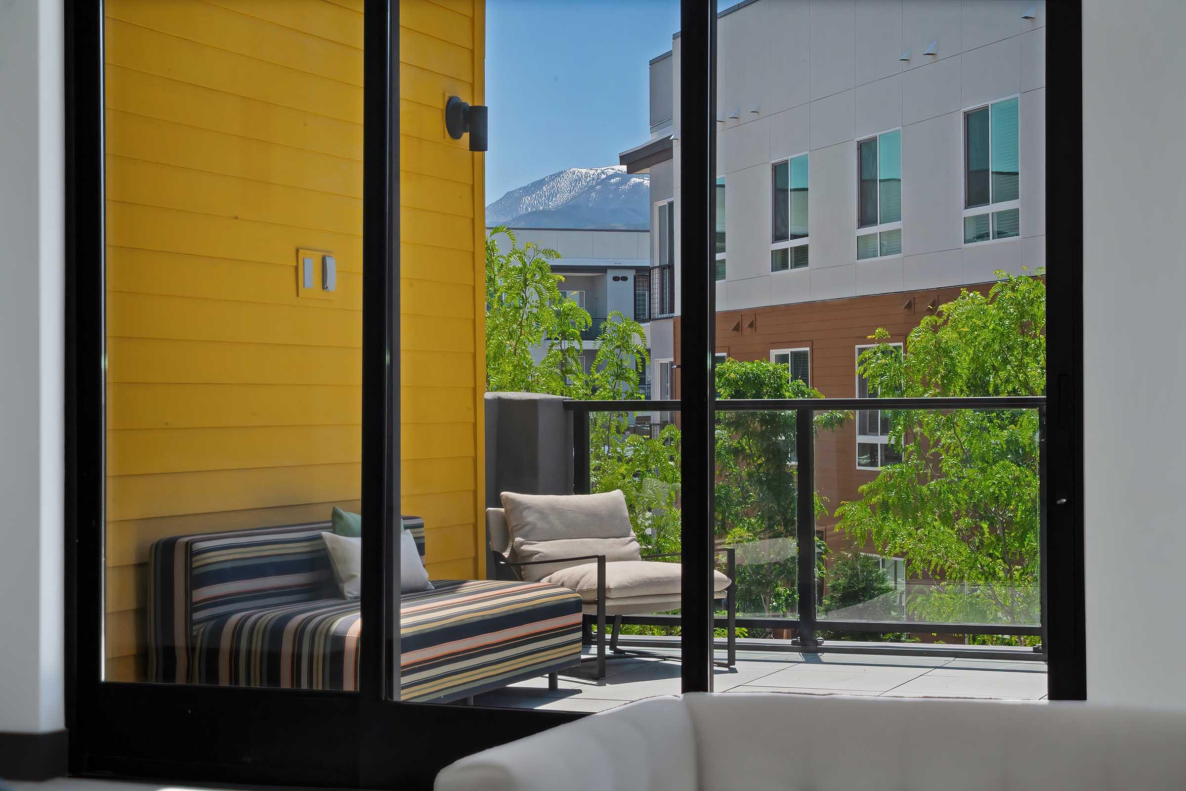 A bright balcony view featuring a striped outdoor couch and a single lounge chair, surrounded by greenery. In the background, modern buildings and a snow-capped mountain are visible under a clear blue sky. The scene conveys a fresh and inviting atmosphere.