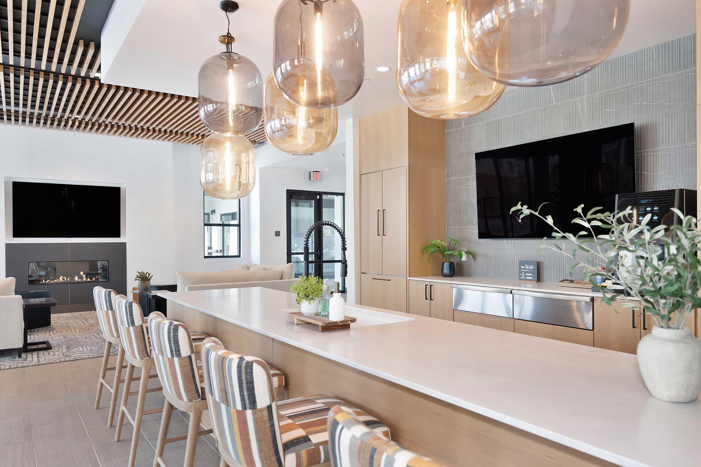 A modern kitchen and lounge area featuring a sleek countertop with bar stools, large decorative pendant lights, a wall-mounted TV, and a stylish backsplash. The space is bright and inviting, with plants and soft furniture that enhance the contemporary design.