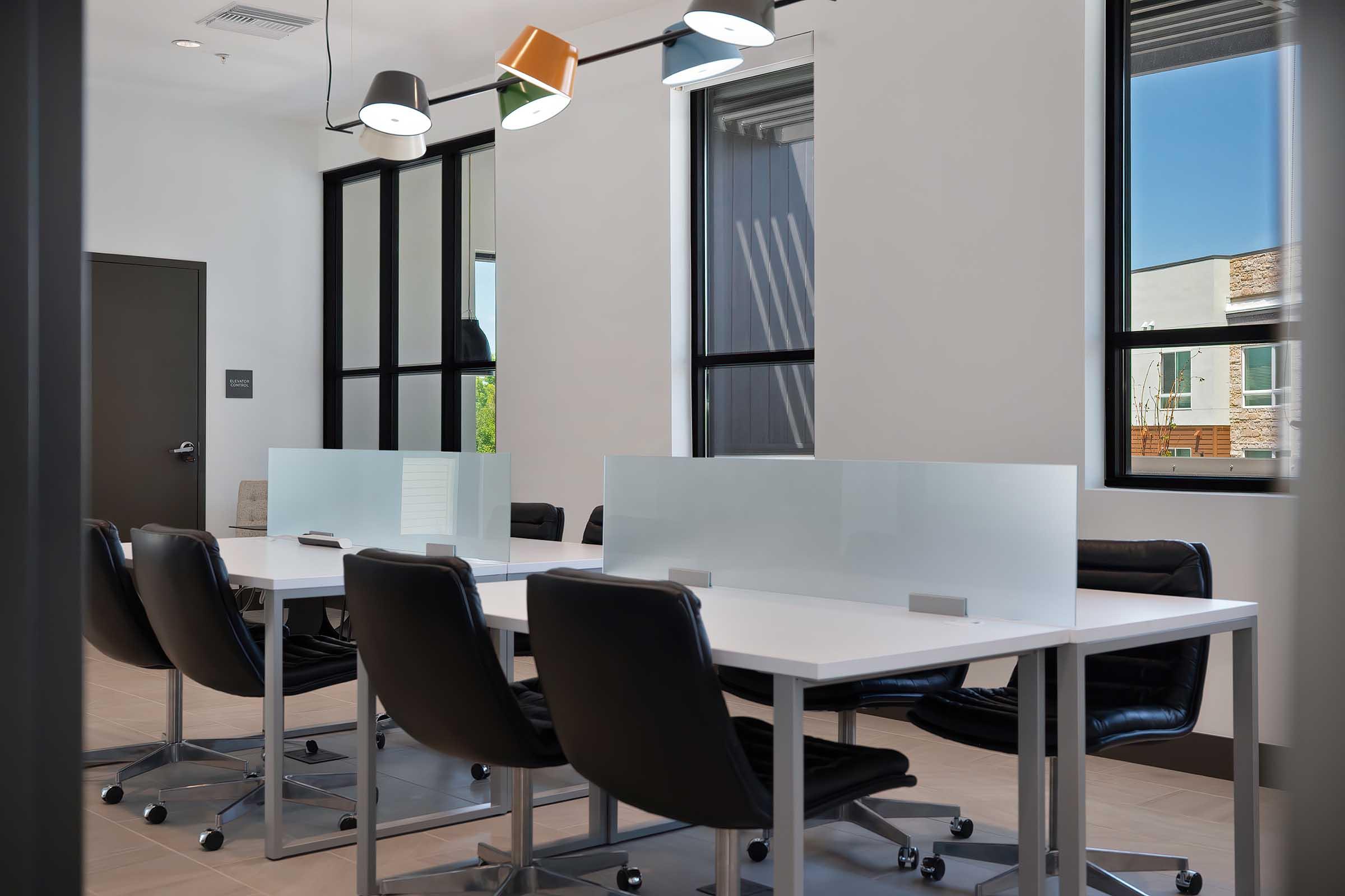 A modern conference room featuring a large white table surrounded by black ergonomic chairs. The room has multiple windows allowing natural light, and sleek pendant lights hang from the ceiling. Glass partitions on the table provide privacy for discussions. Minimalistic decor enhances the professional atmosphere.
