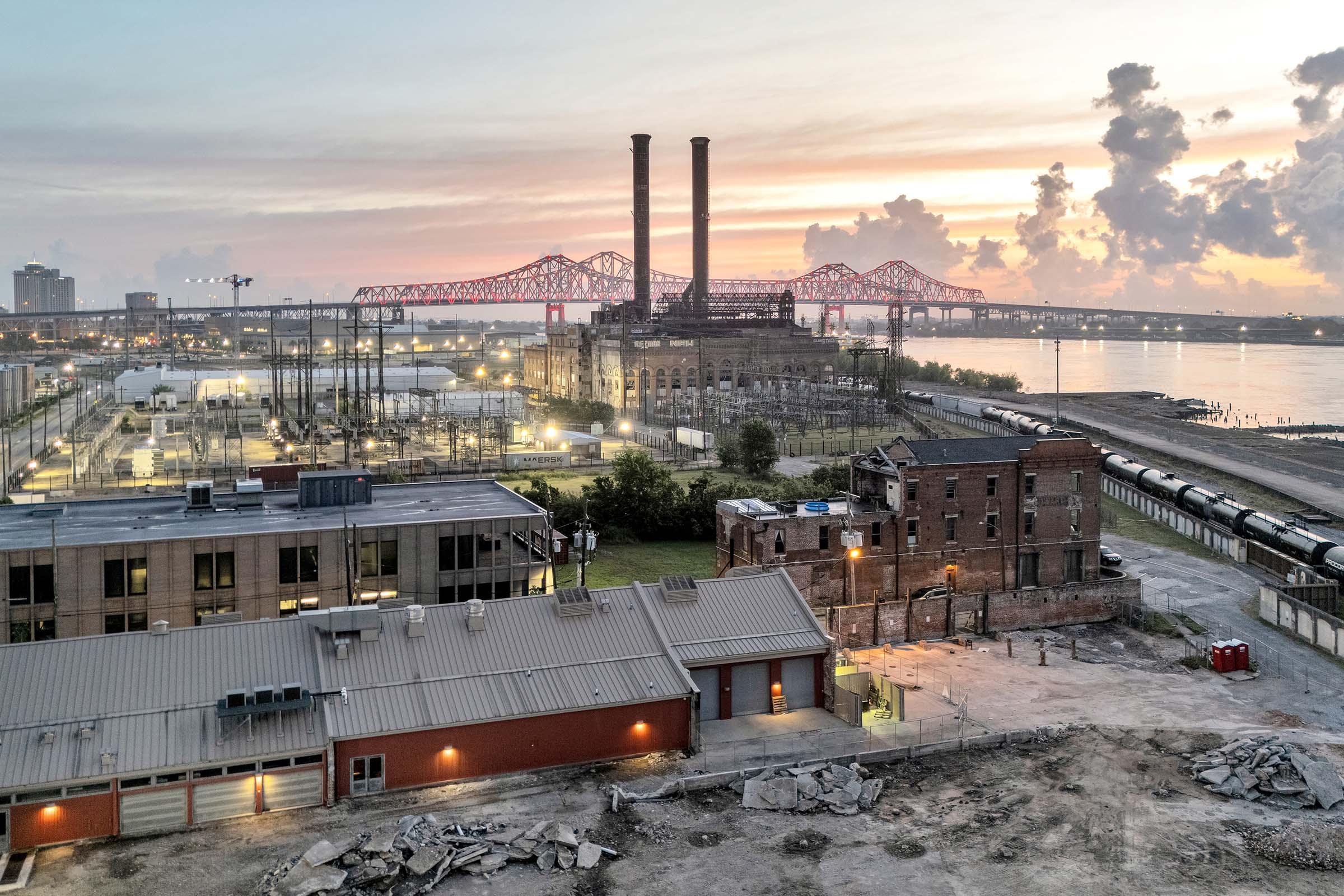 Aerial view of a historic industrial site at sunset, featuring two tall smokestacks and a partially demolished brick building. The scene includes nearby structures, power lines, and a bridge in the background, with the river visible and lit by surrounding lights. The sky is filled with pastel colors as day transitions to night.