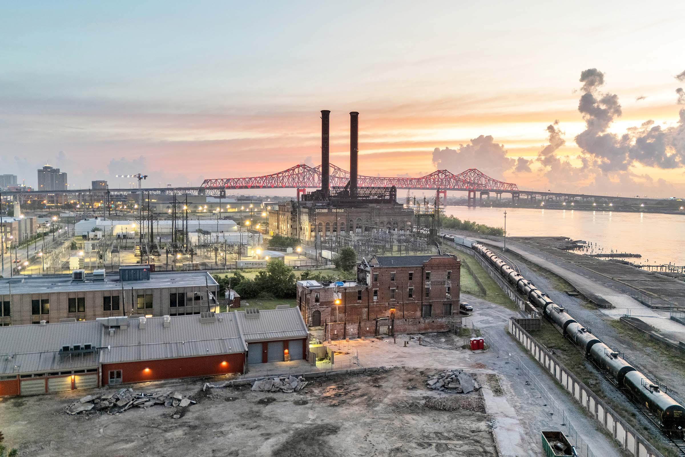 Aerial view of a city industrial area at sunset, featuring a historic power plant with two tall smokestacks, surrounded by buildings and rail tracks. The Mississippi River is visible in the background, along with a bridge illuminated by the sunset, casting a warm glow over the scene.