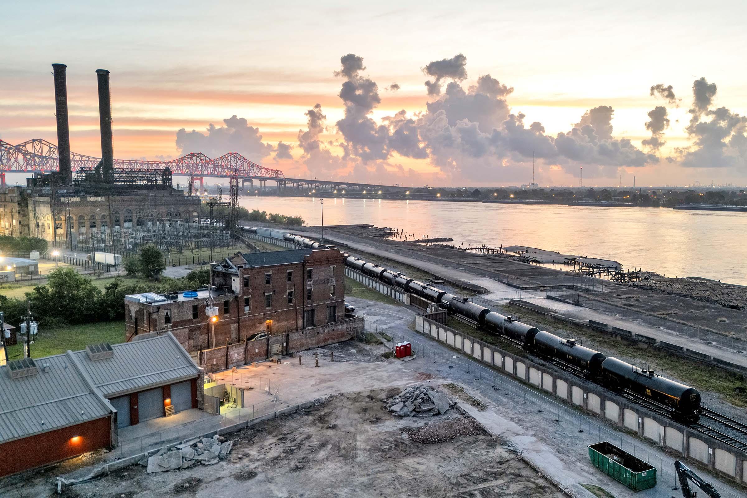 A scenic view of an industrial landscape at sunset, featuring a river, a train with tank cars, and a historic building. Smokestacks rise in the background amidst a colorful sky with clouds. The environment appears to be undergoing development, with construction materials visible on the ground.
