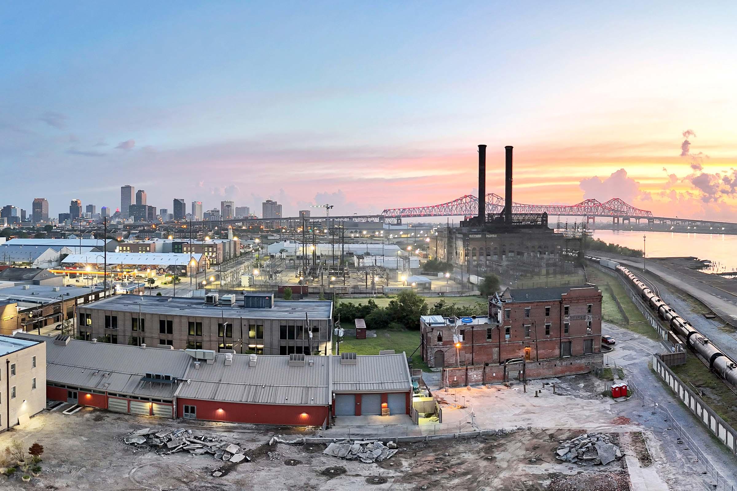 A panoramic view of a city skyline at sunset, featuring a river and industrial buildings in the foreground. Notable structures include a historic smokestack and a bridge in the distance. The sky is filled with pastel colors as a train line runs along the edge of the image, highlighting urban renewal elements.