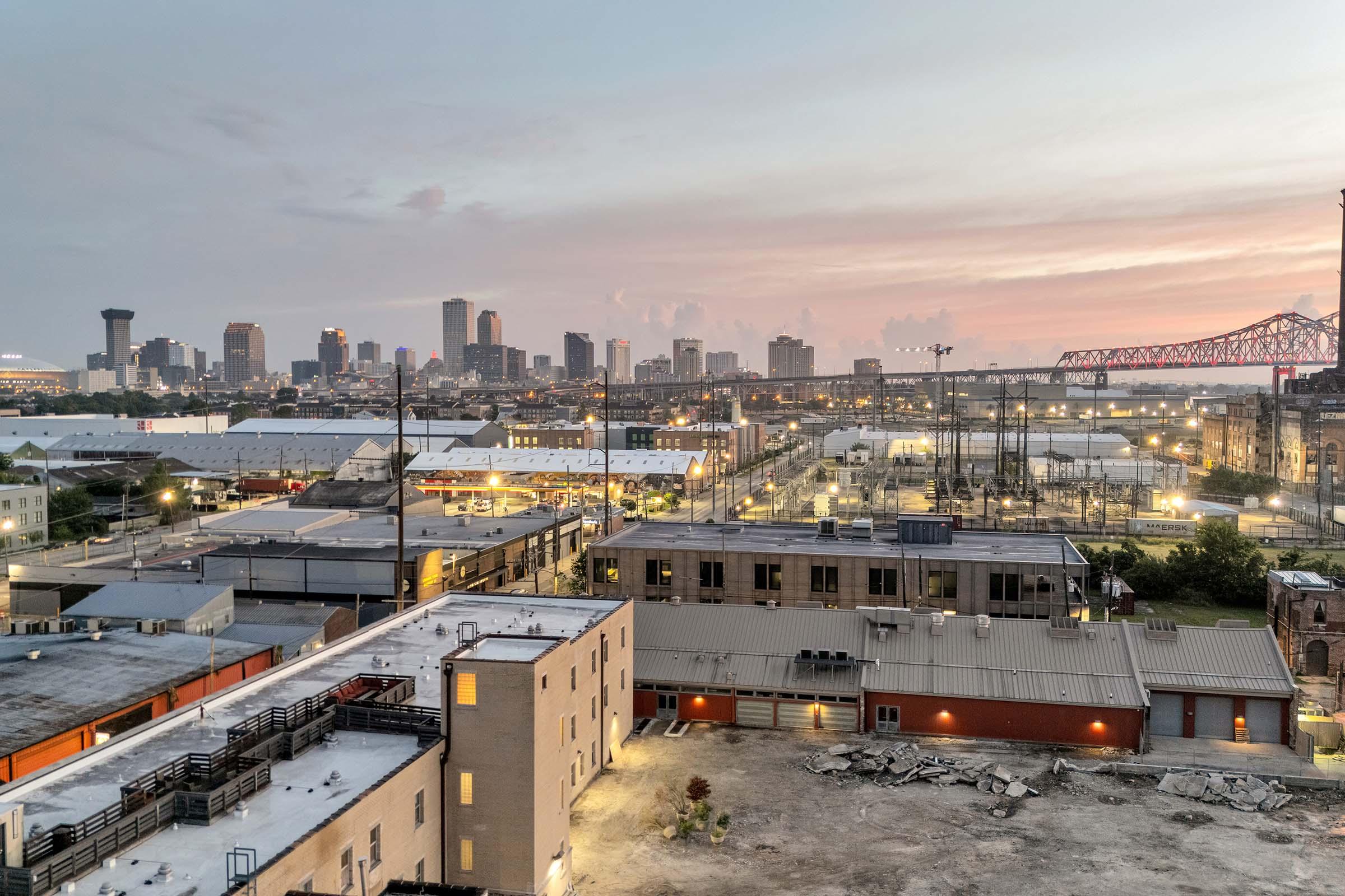 A panoramic view of a city skyline at dusk, featuring tall buildings and a bridge in the background. The foreground shows industrial buildings and some construction debris. The sky has soft hues of pink and orange as the sun sets, illuminating the urban landscape.