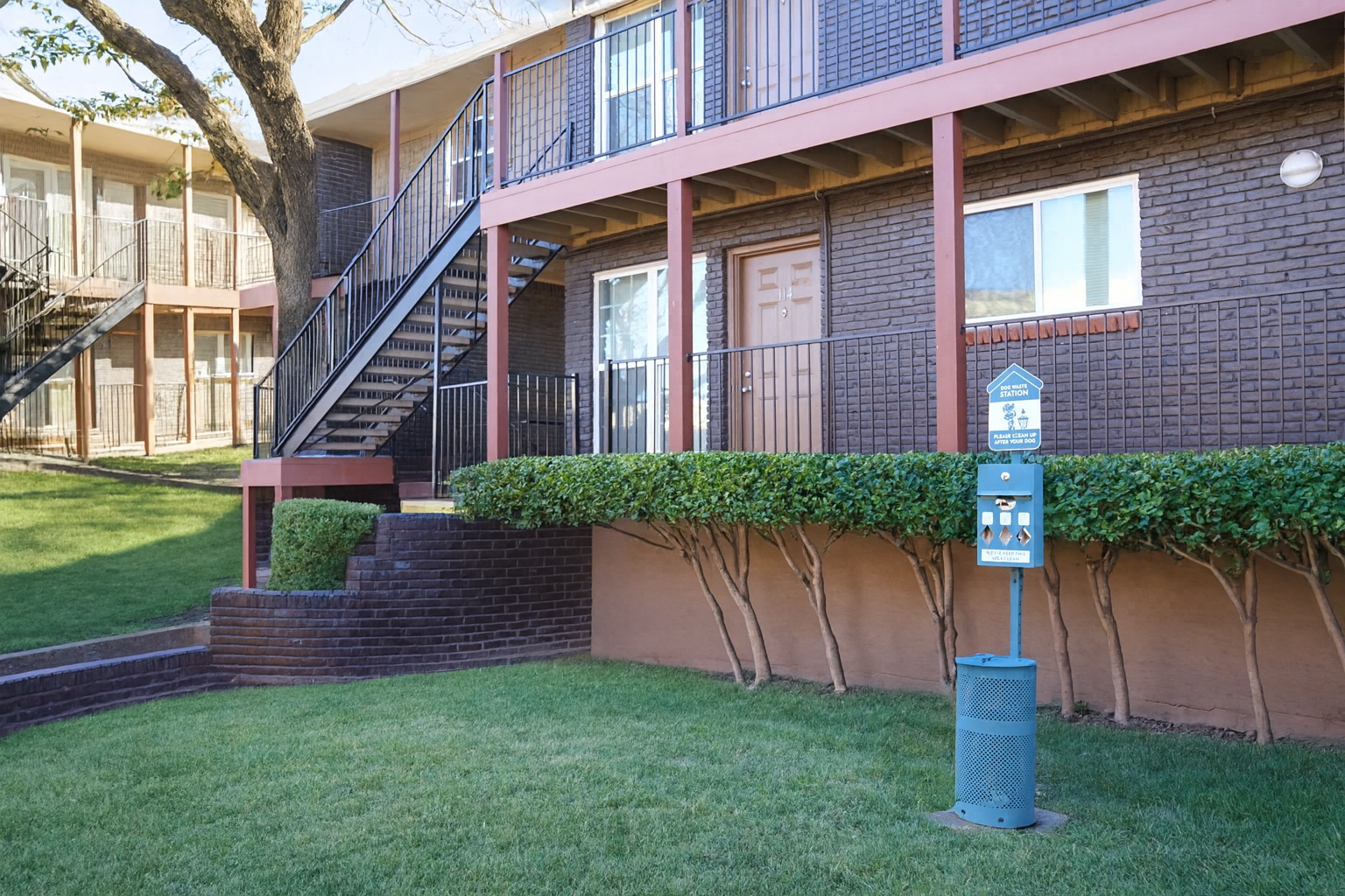 A multi-story apartment complex with a low brick wall, neatly trimmed hedges, and a grassy area. There are stairs leading up to a balcony on the left side, and a sign next to a pet waste station is visible on the right. The building features a brown exterior with several windows.
