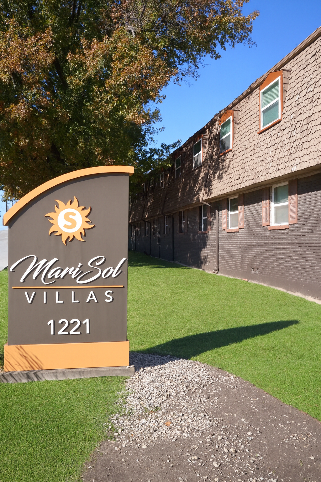 Sign for MariSol Villas with the address 1221, in front of a building. The sign features a sun icon and is set against a backdrop of green grass and trees. The building has a shingled roof and windows with brown trim.