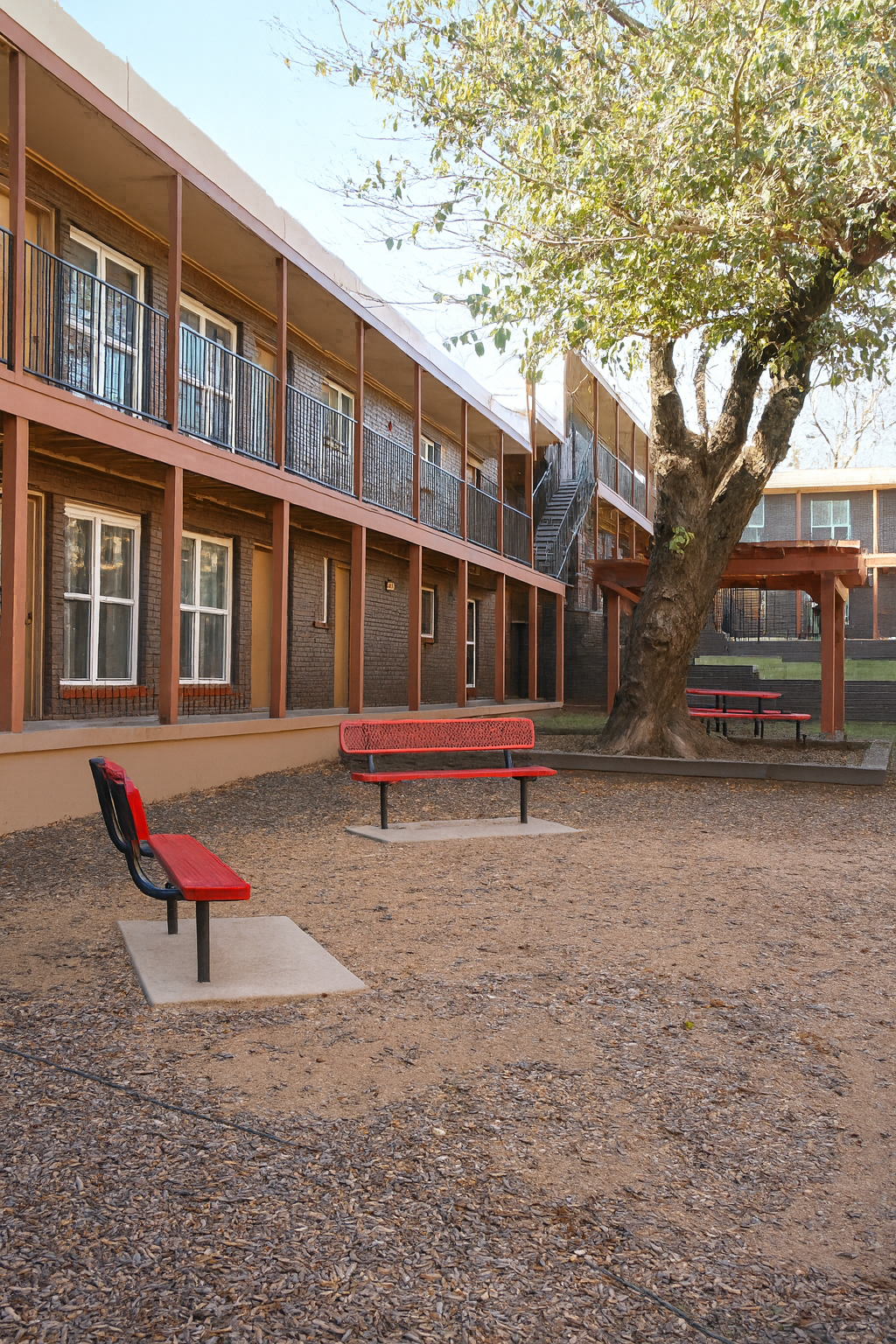 A courtyard area with a red bench and a matching chair surrounded by gravel. In the background, there are two-story buildings with balconies and windows. A large tree provides shade to part of the space, which also features another seating area with picnic tables. The scene is bright and open, conveying a welcoming environment.