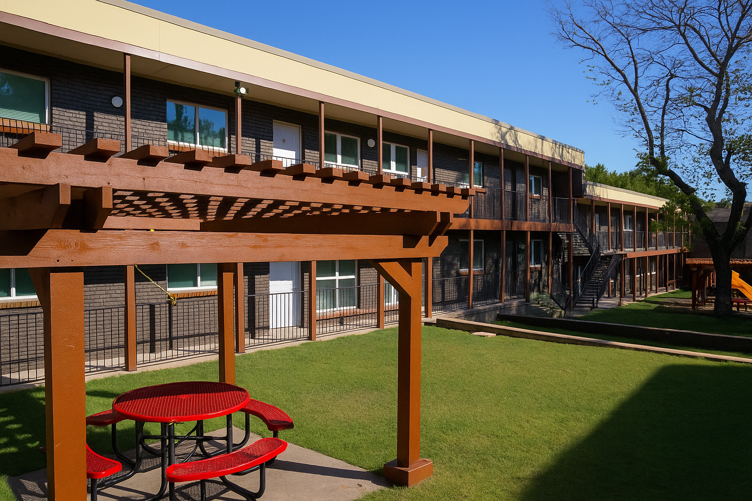 A sunny courtyard of a multi-story building, featuring a wooden pergola with a picnic table underneath. The green lawn is well-kept, and there are balconies on the upper floor. In the background, a tree adds natural beauty to the scene, with blue skies overhead.