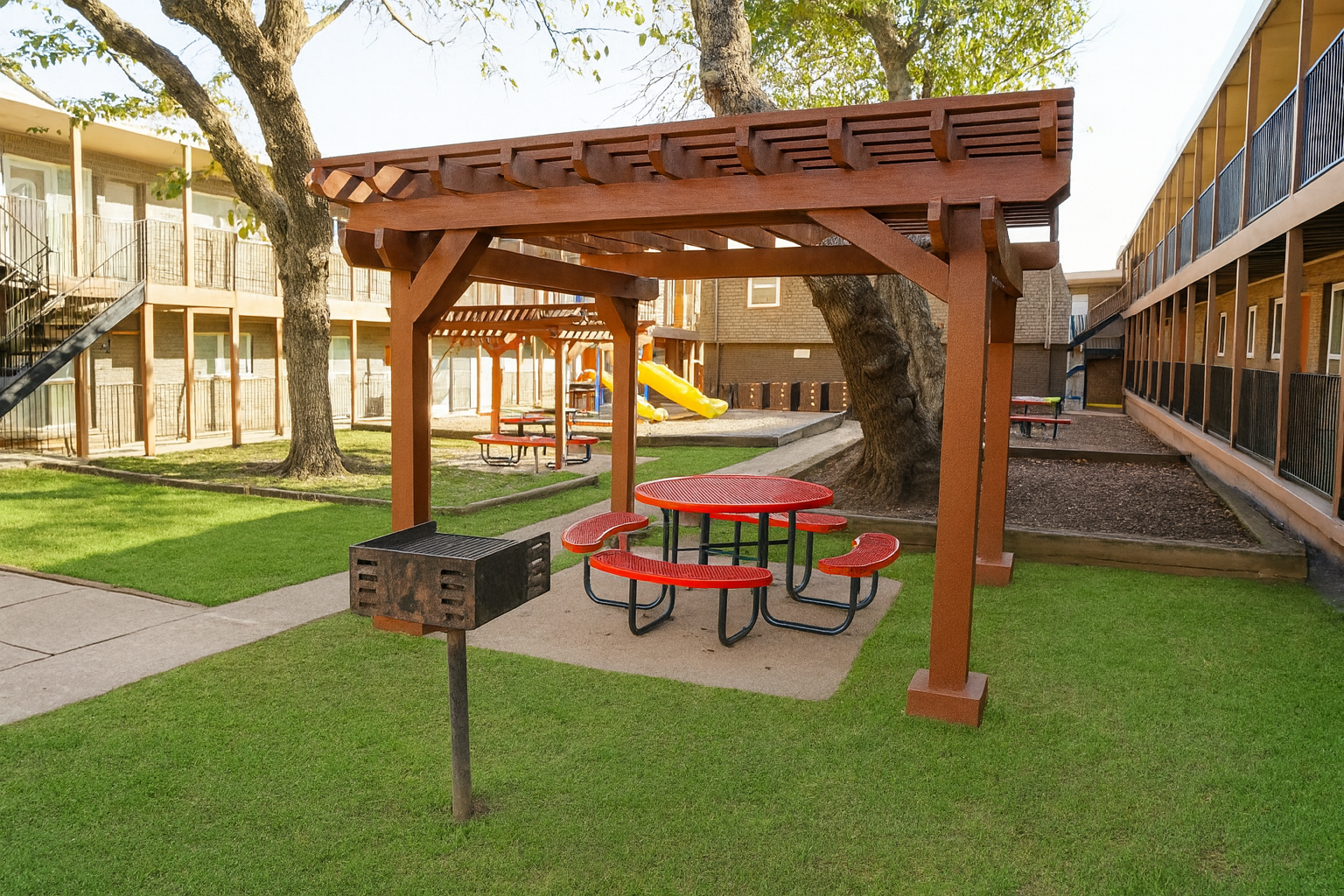 A wooden pergola with red picnic tables underneath sits on a green lawn. Nearby, a grill is positioned next to the seating area. In the background, there are two-story buildings with balconies and a playground visible. The scene is well-lit, suggesting a bright day.