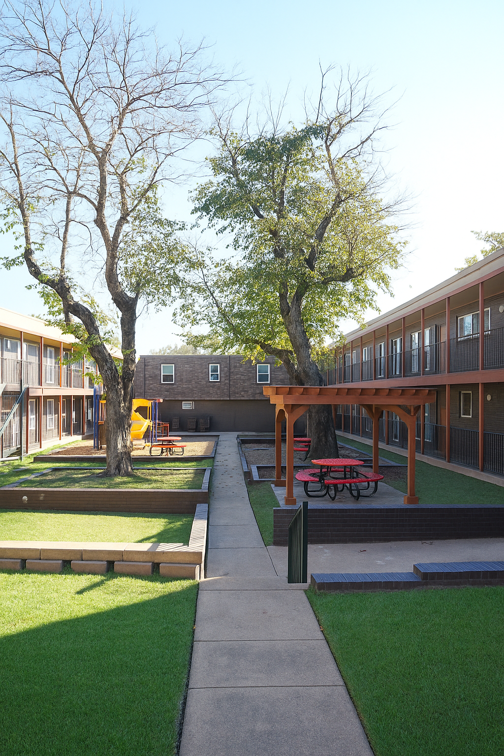 A courtyard area surrounded by two-story apartment buildings. It features grassy sections, a paved walkway, a playground with slides, and picnic tables under a wooden pergola. Several trees are visible, and the scene is set in bright, clear weather.