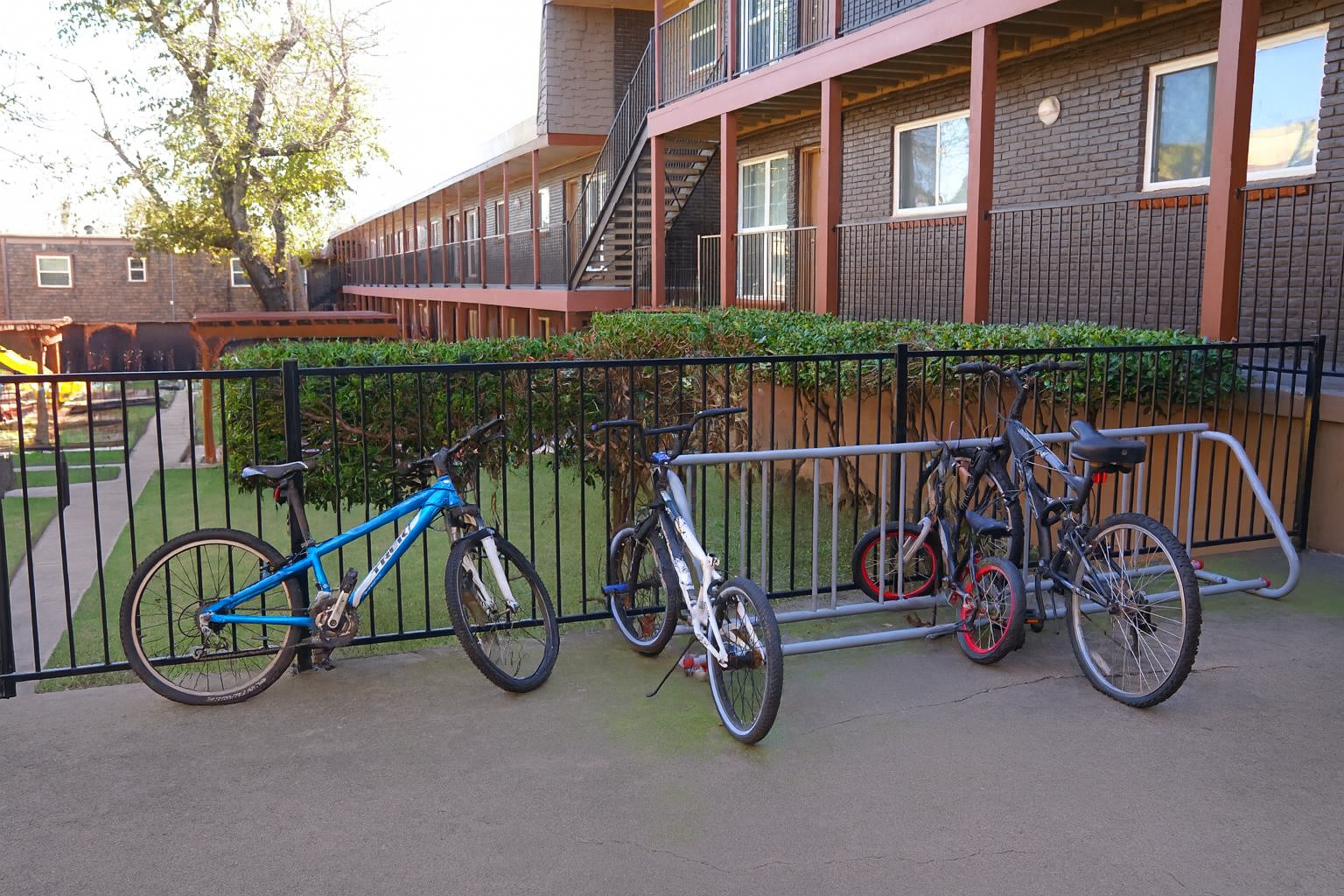 A row of bicycles parked next to a fence in a courtyard area of an apartment complex. The scene includes various types of bikes, with a backdrop of greenery and the building's architecture.