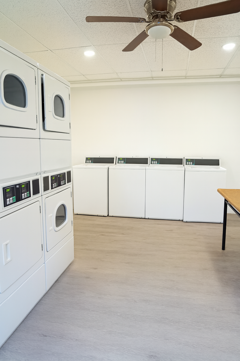 A bright laundry room featuring stacked washing machines and dryers on the left, and several front-loading machines along the back wall. A wooden table is positioned on the right, and a ceiling fan is visible above, providing a clean and functional space for laundry activities.