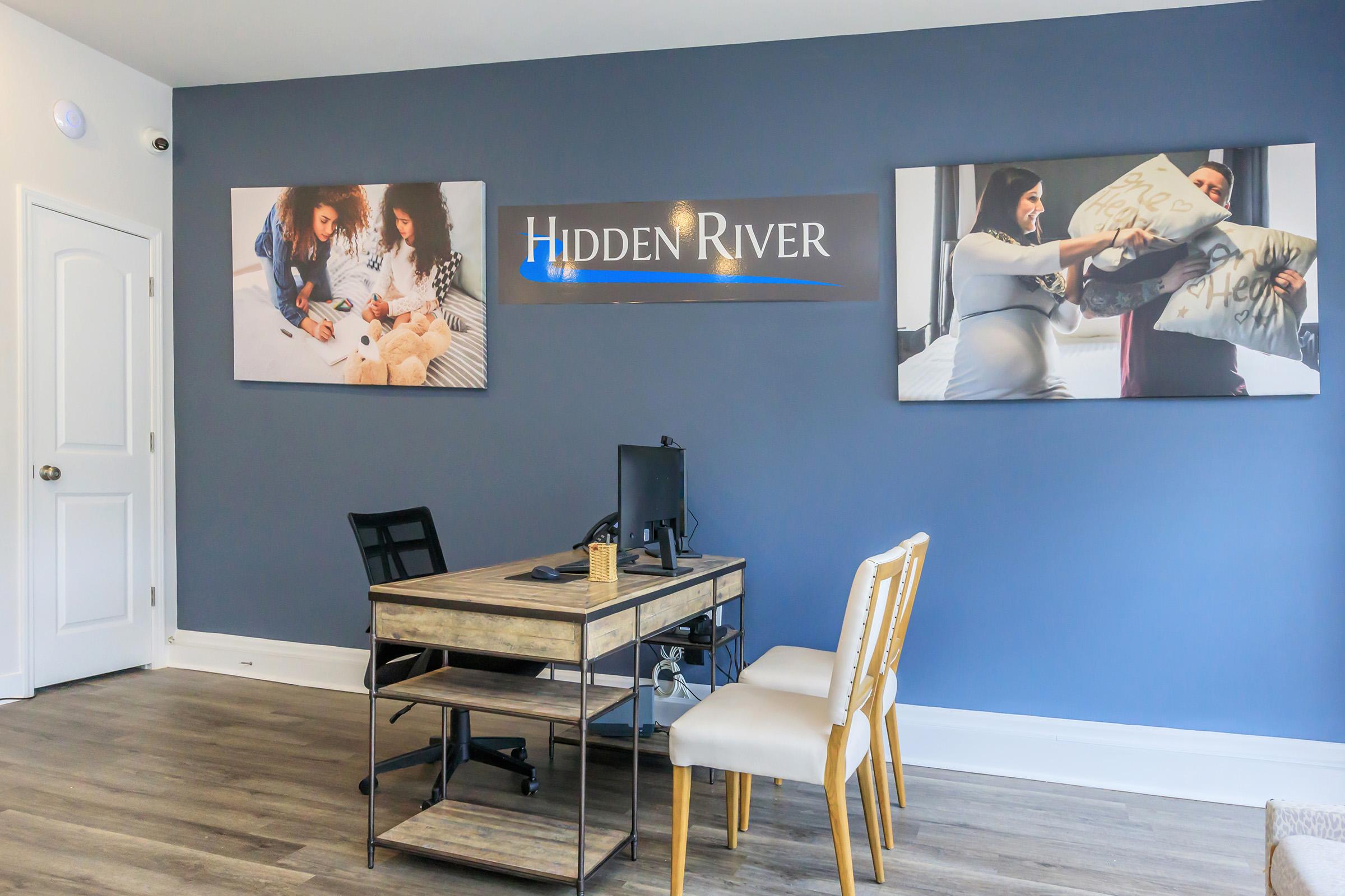 A modern office interior featuring a wooden desk with a computer, two chairs, and wall art. The grey wall displays the text "Hidden River" and two photographs: one depicting a family with children and another showing a couple on a couch. The flooring is light-colored wood.
