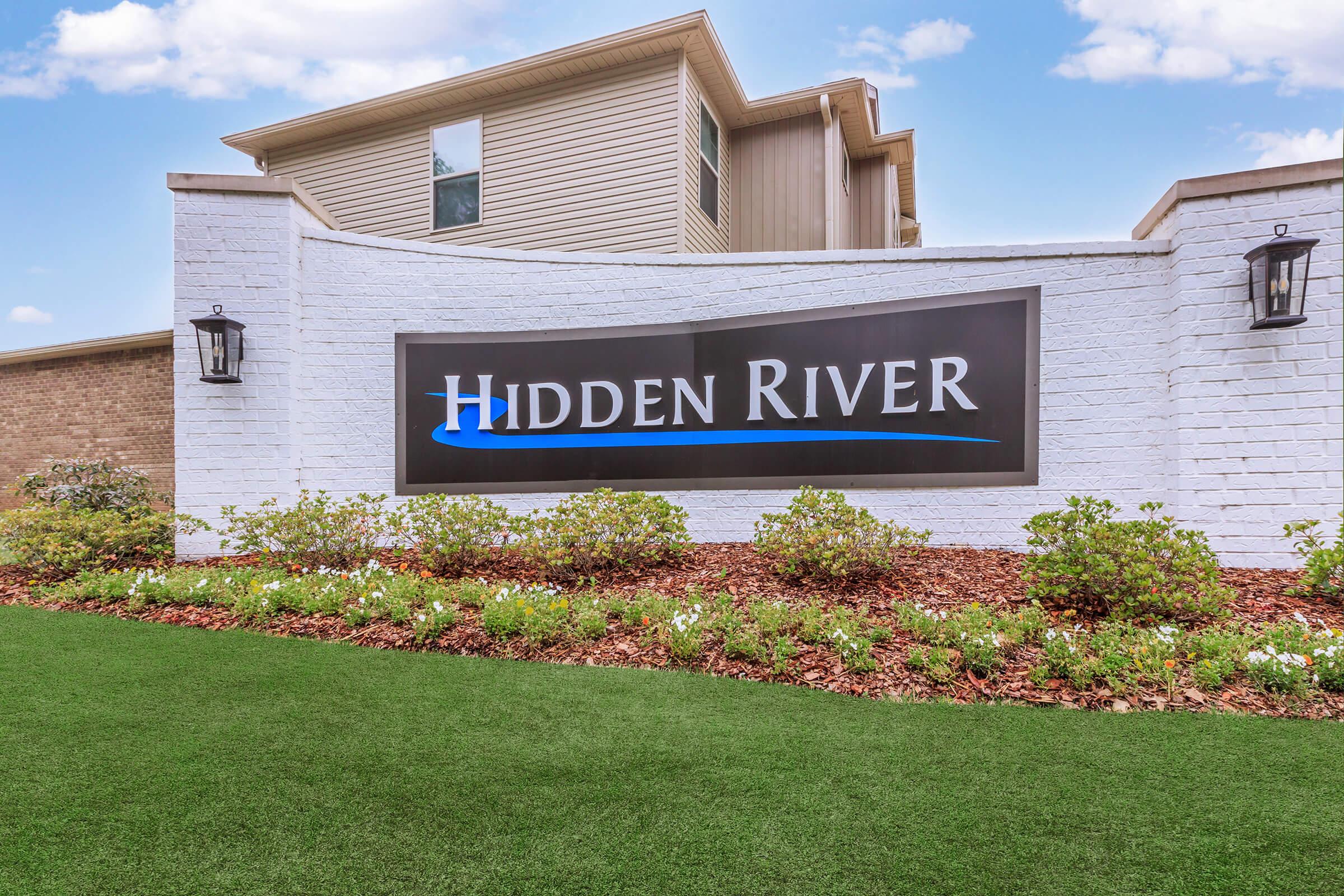 Sign for Hidden River, featuring the name prominently displayed in blue text on a black background. The sign is set against a white brick wall, surrounded by landscaped greenery and flowers, with a building visible in the background. Lush green grass is in the foreground.