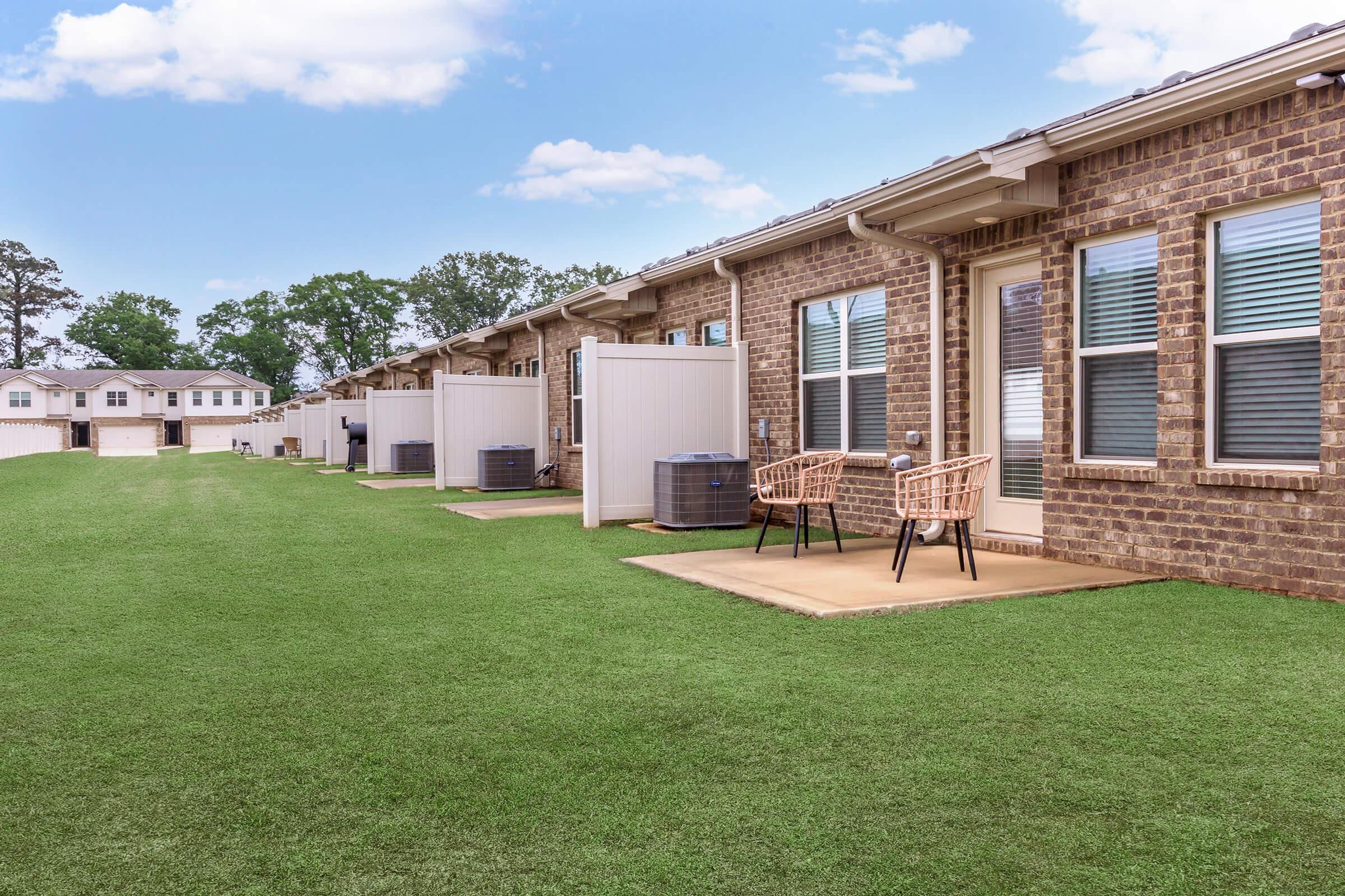 Row of modern brick apartments with small patios, surrounded by well-maintained green grass. Each unit features large windows and air conditioning units, with a few wicker chairs placed outside for seating. A clear blue sky with scattered clouds creates a bright atmosphere.