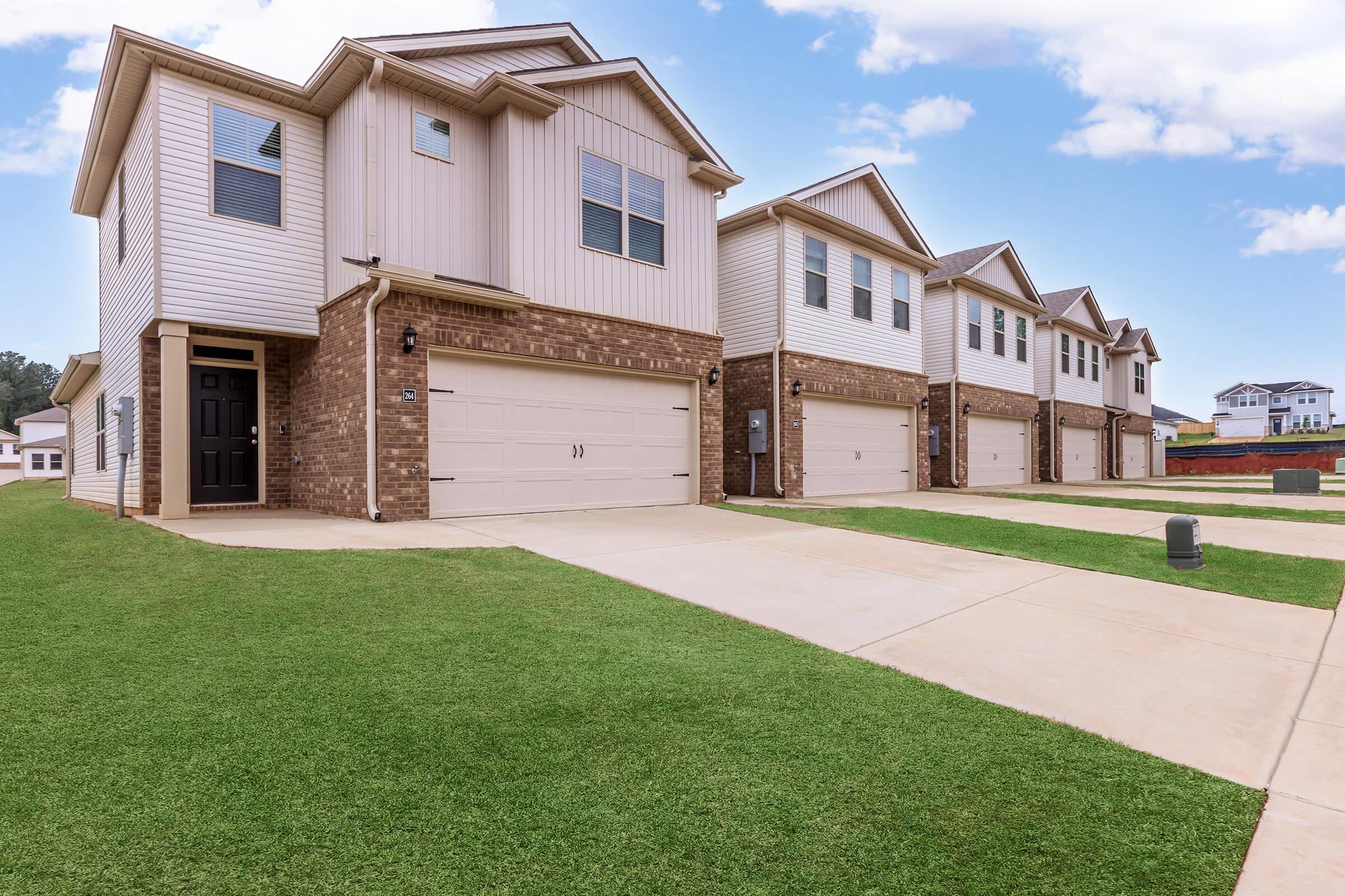 A row of modern two-story homes with brick and siding exteriors, featuring garages and well-maintained lawns. The sky is partly cloudy, highlighting the clean and inviting neighborhood setting. Each home has multiple windows and a symmetrical design, creating a cohesive appearance.