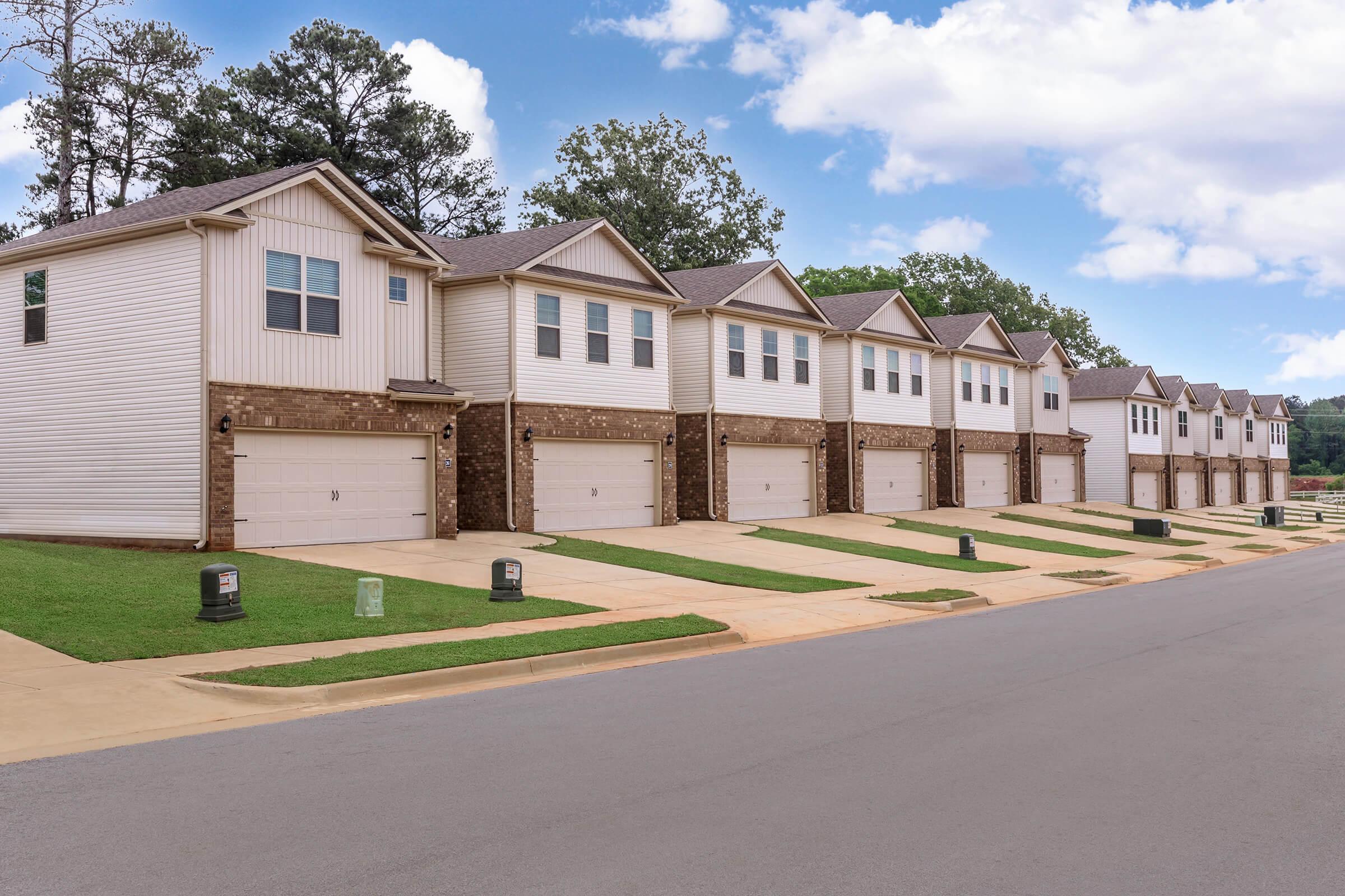 Row of modern townhouses with garages, featuring beige siding and stone accents, set against a clear blue sky. Neatly landscaped lawns with green grass line the driveways, and trash bins are visible in front of some units. Trees are present in the background, adding to the suburban setting.