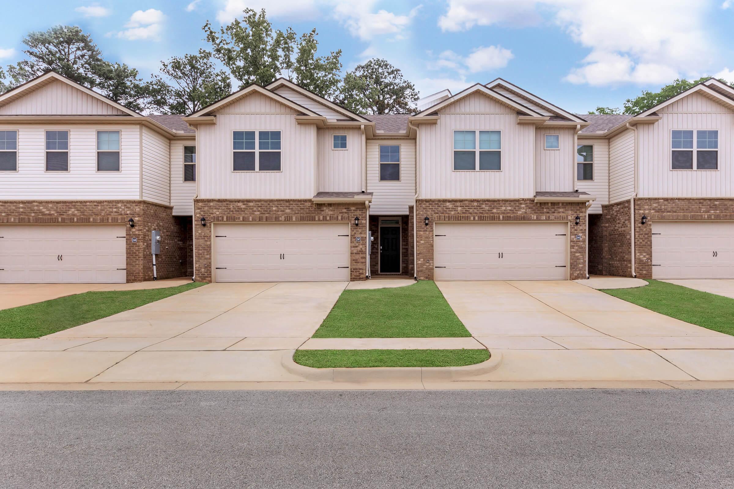 Row of modern townhouses featuring brick and siding exteriors, each with a two-car garage. The driveways are paved, and there is well-maintained green grass in front of the homes. The sky is partly cloudy, creating a pleasant atmosphere.