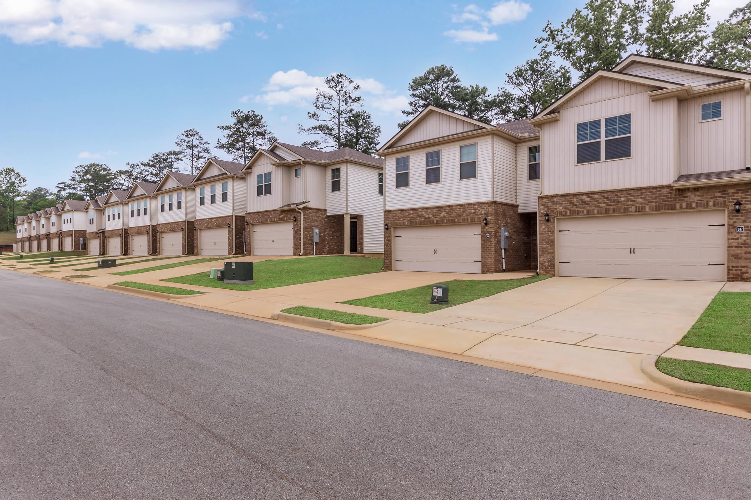 A quiet residential street featuring a row of modern townhouses. Each townhouse has a two-car garage and is surrounded by well-maintained grass. The houses are lined up neatly along the street, with trees visible in the background under a partly cloudy sky.