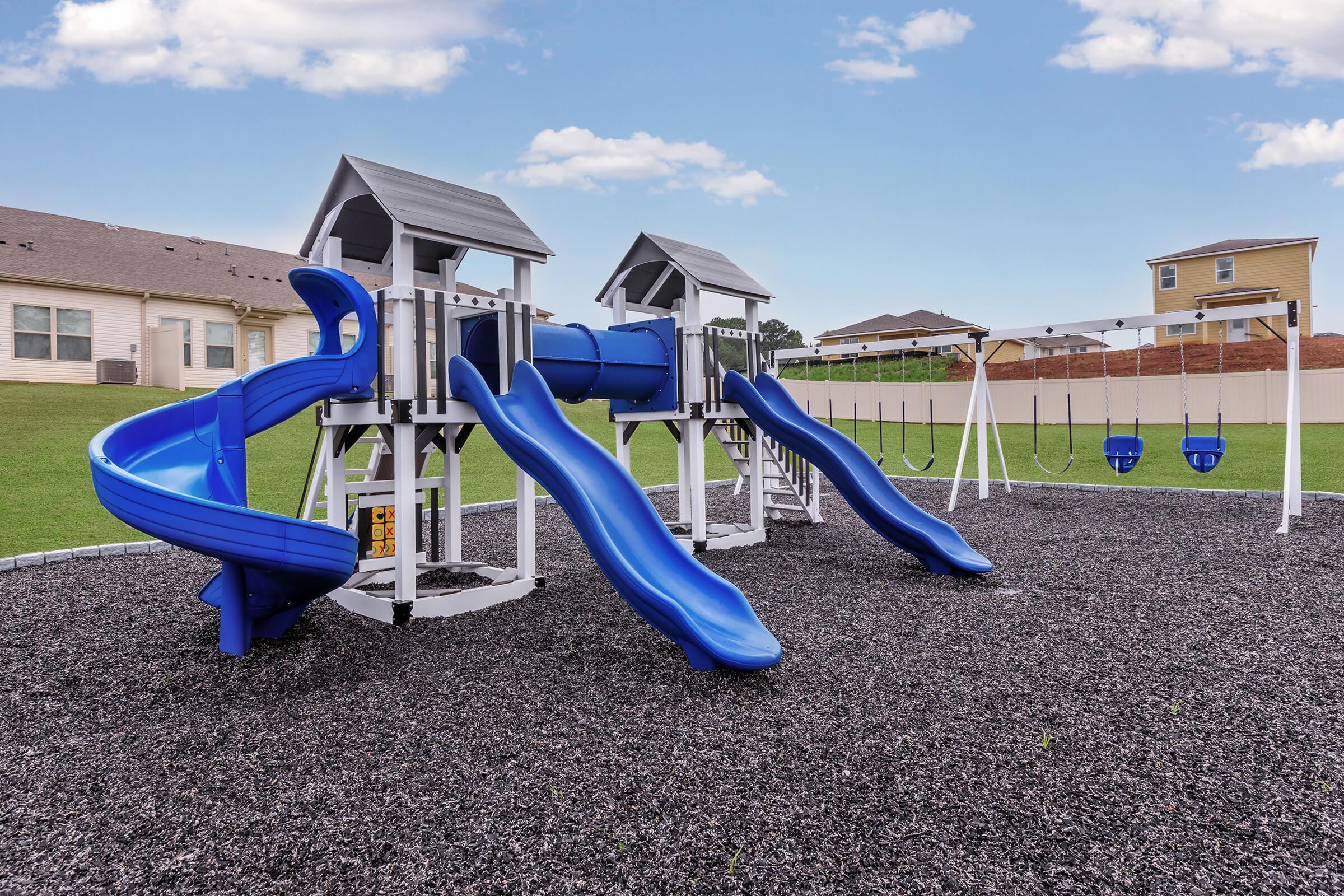 Playground equipment featuring two tall blue slides, a climbing structure with two roofed lookout towers, and a swing set with two swings, all situated on a gravel surface. The background shows a grassy area and residential buildings under a clear blue sky.