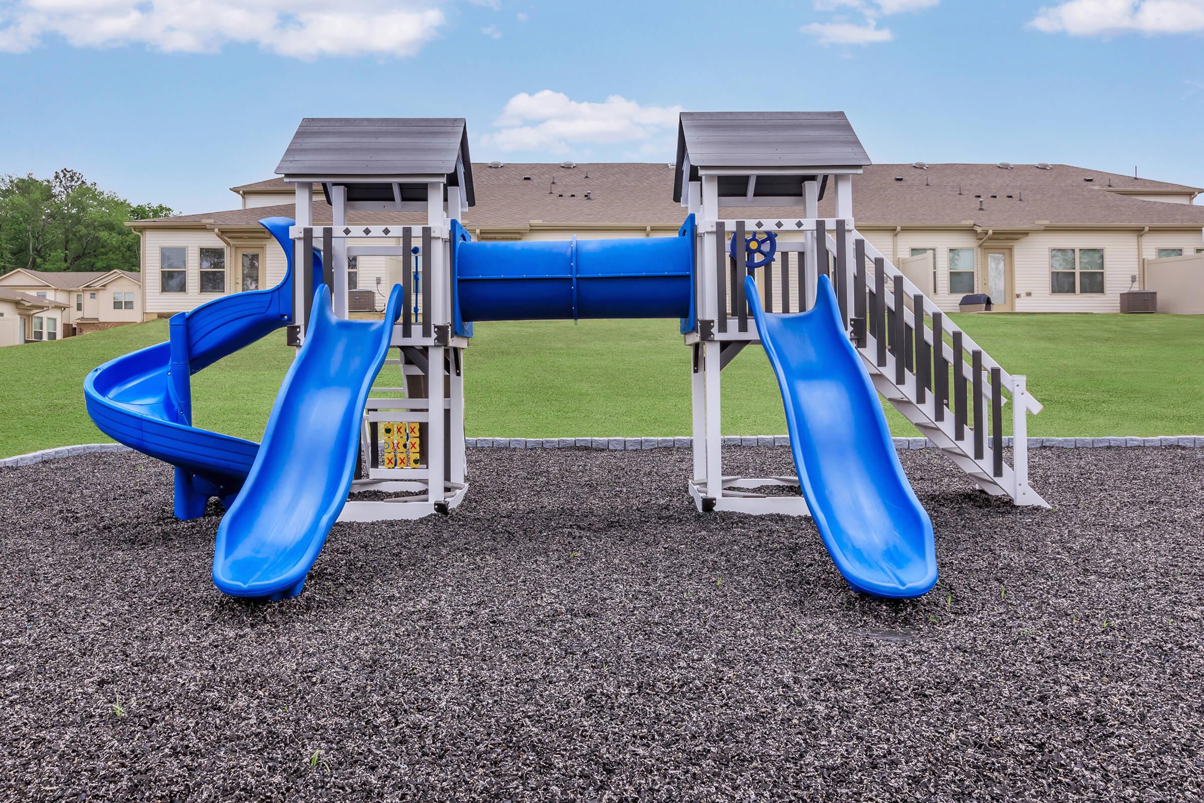 A playground structure featuring two blue slides, a tunnel, and two playhouses. The play area is surrounded by black mulch and backed by grassy fields and residential buildings under a clear blue sky.