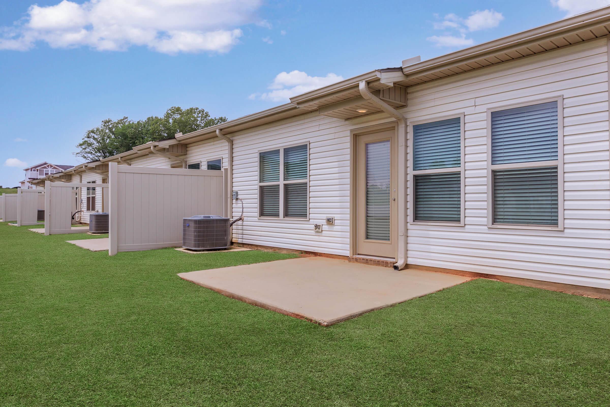 A side view of a single-story residential building with a light-colored exterior. The building features several windows and a door, surrounded by a well-maintained lawn with artificial grass. There are air conditioning units visible on one side, and a privacy fence separates the property from the neighboring area.