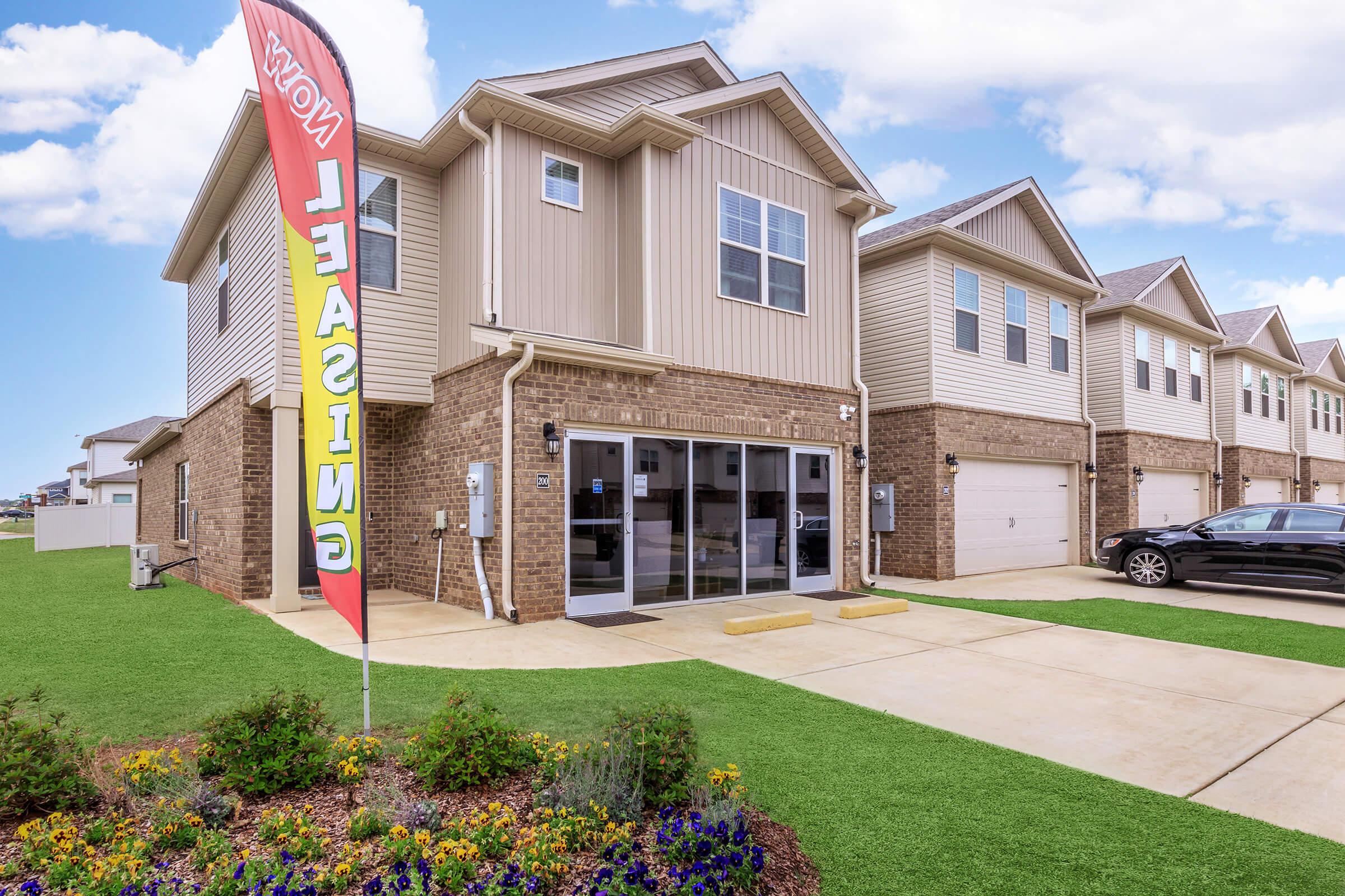 A modern, two-story residential building with beige siding and brick accents. The front features large windows and a glass door, with a colorful "Now Leasing" banner displayed. A well-maintained lawn with blooming flowers is in the foreground, and a parked car is visible in the driveway.