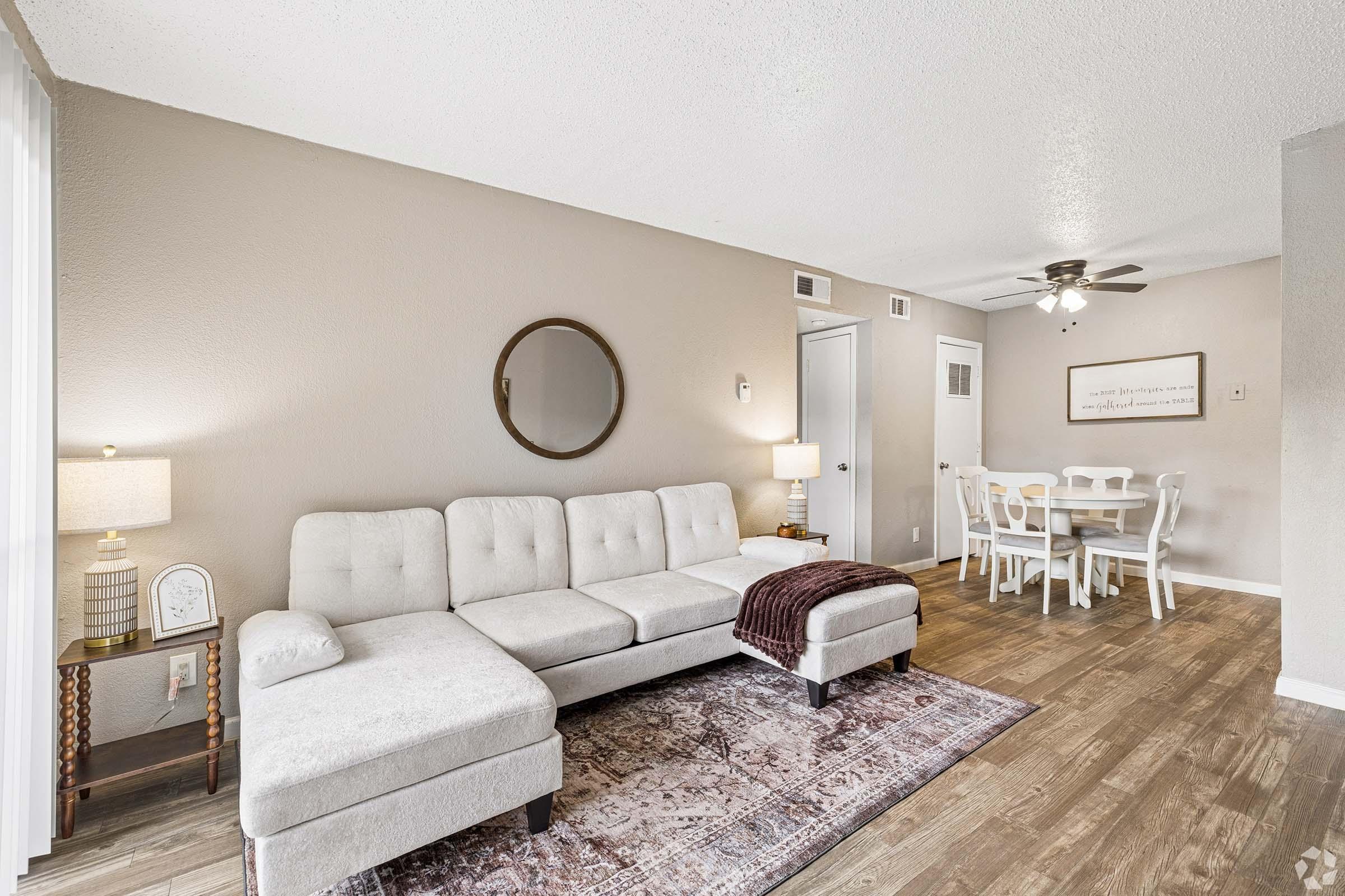A cozy living room featuring a light gray sectional sofa with a maroon throw, a round mirror on the wall, a lamp on a side table, and a patterned area rug. In the background, a dining area with a white table and chairs is visible, along with a ceiling fan and neutral-colored walls.