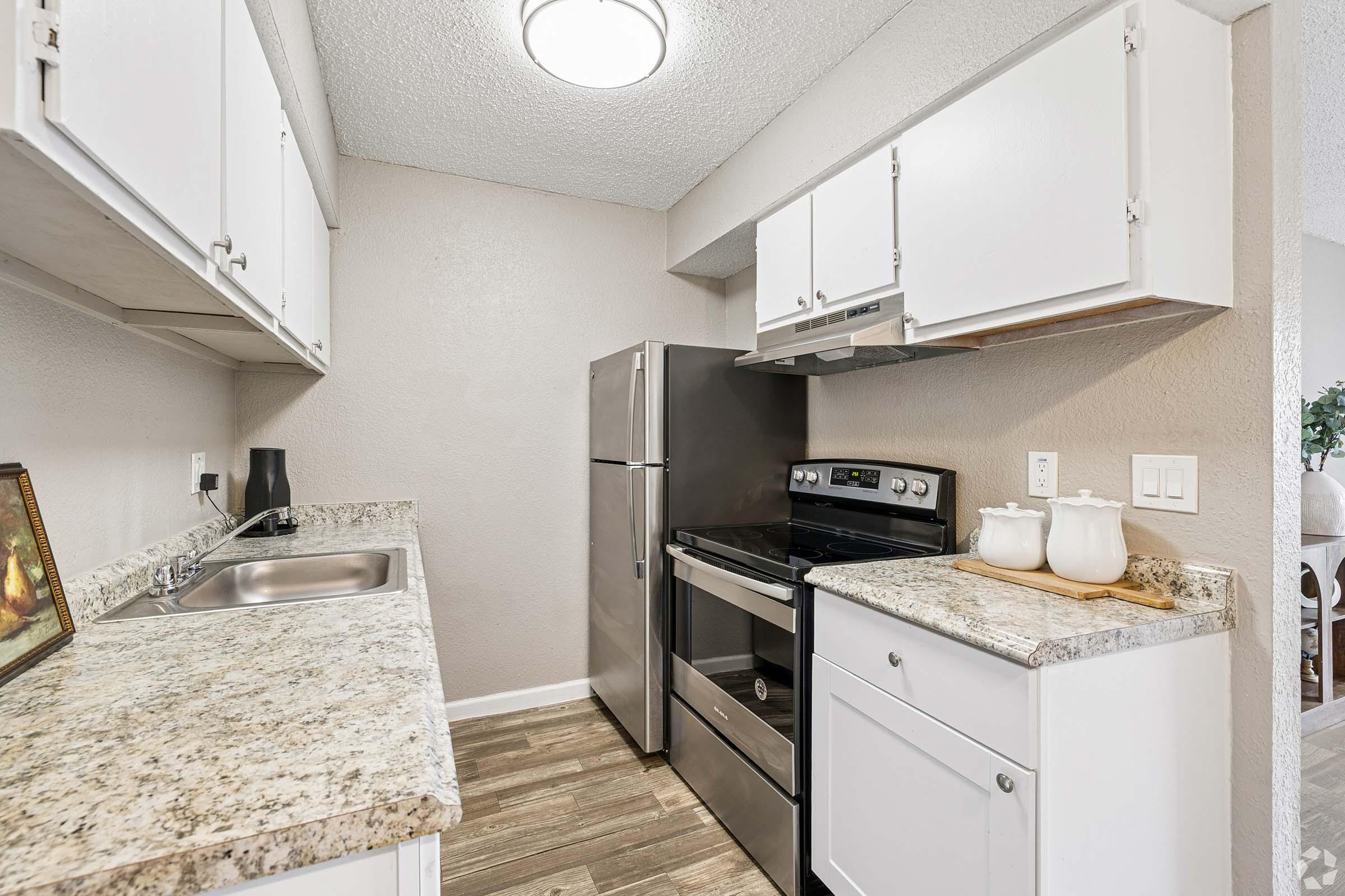 Bright kitchen featuring white cabinets, a granite countertop, and stainless steel appliances, including a refrigerator and stove. The space is well-lit with a ceiling light, and the floor has a wood-like finish. A small sink is visible, and there is a decorative picture on the counter.