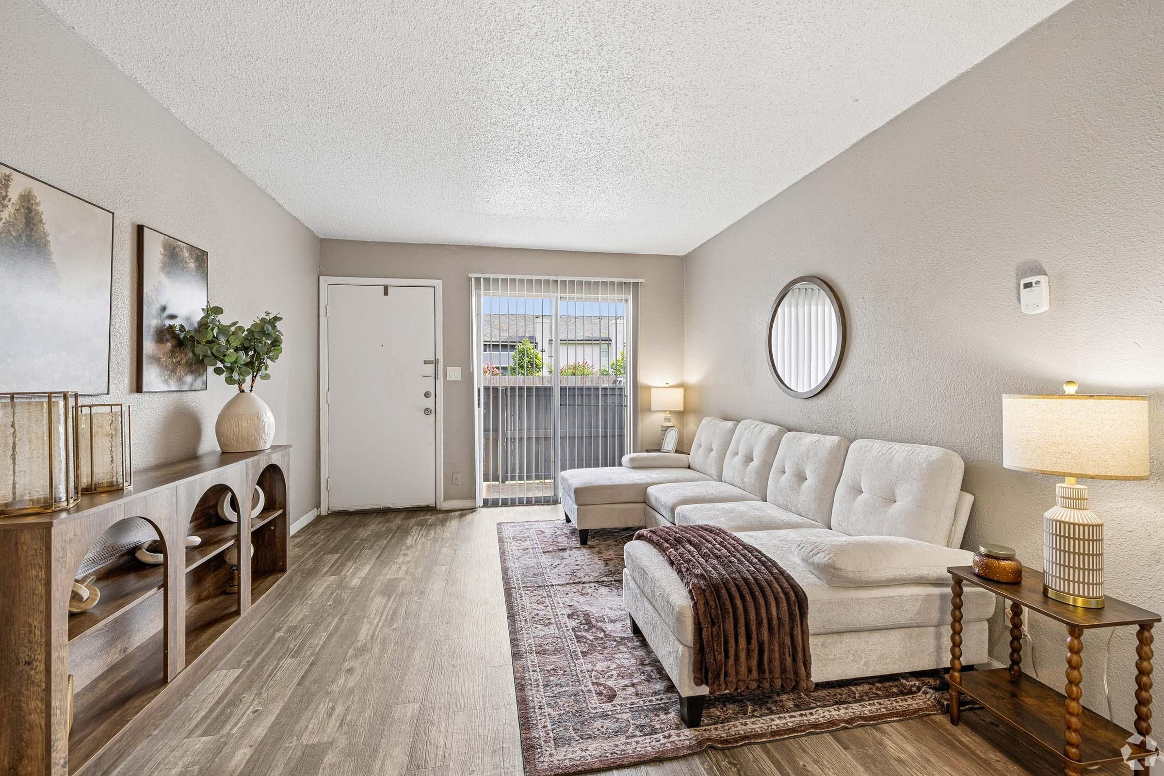 A cozy living room featuring a light-colored sectional sofa with a brown throw blanket, a wooden console table, and a round mirror on the wall. There are decorative items on the table, a lamp, and a door leading outside. Natural light filters in through a window, creating a warm and inviting atmosphere.