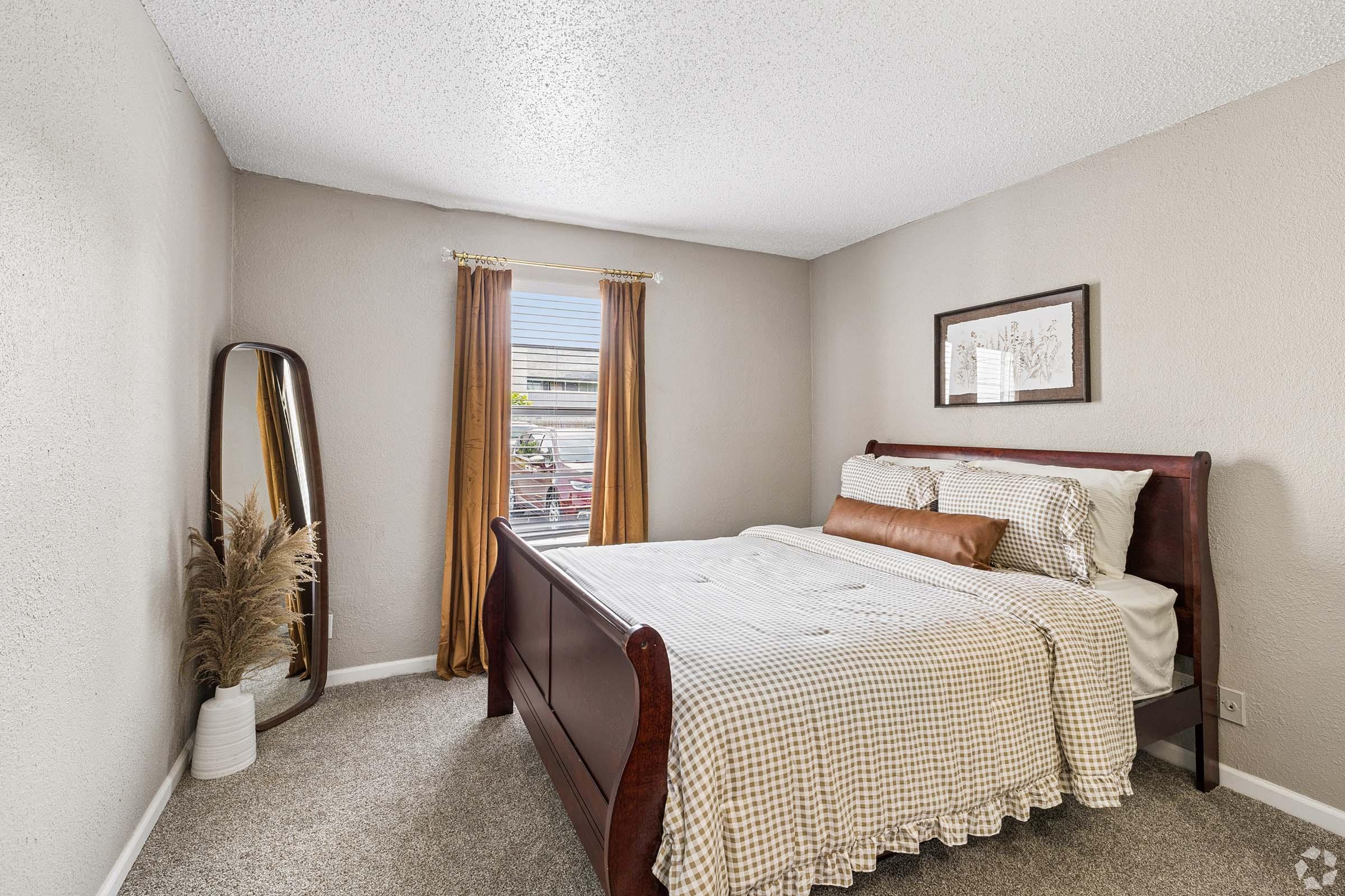 A cozy bedroom featuring a wooden bed with a plaid duvet, a decorative pillow, and a piece of wall art. Natural light filters through a window with curtains, illuminating a mirror and a potted plant in the corner. The walls are painted in a soft hue, and the carpeted floor adds warmth to the space.