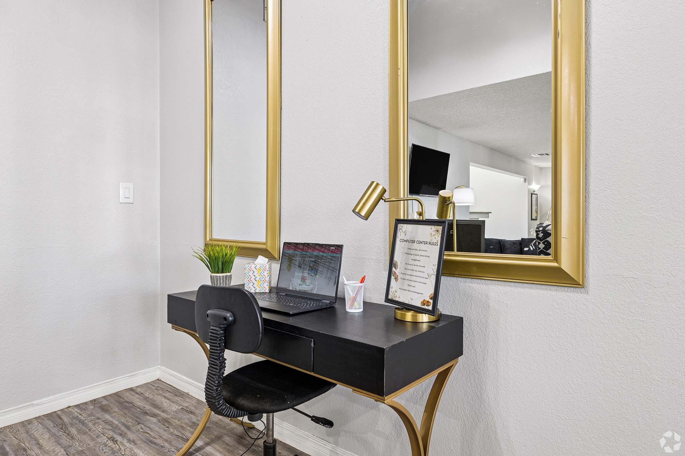 A modern workspace featuring a sleek black desk with a laptop, a chair, and decorative items. Two large gold-framed mirrors are hung above the desk, reflecting the light and creating a spacious feel. A small plant and a framed document add a touch of personalization to the setup.