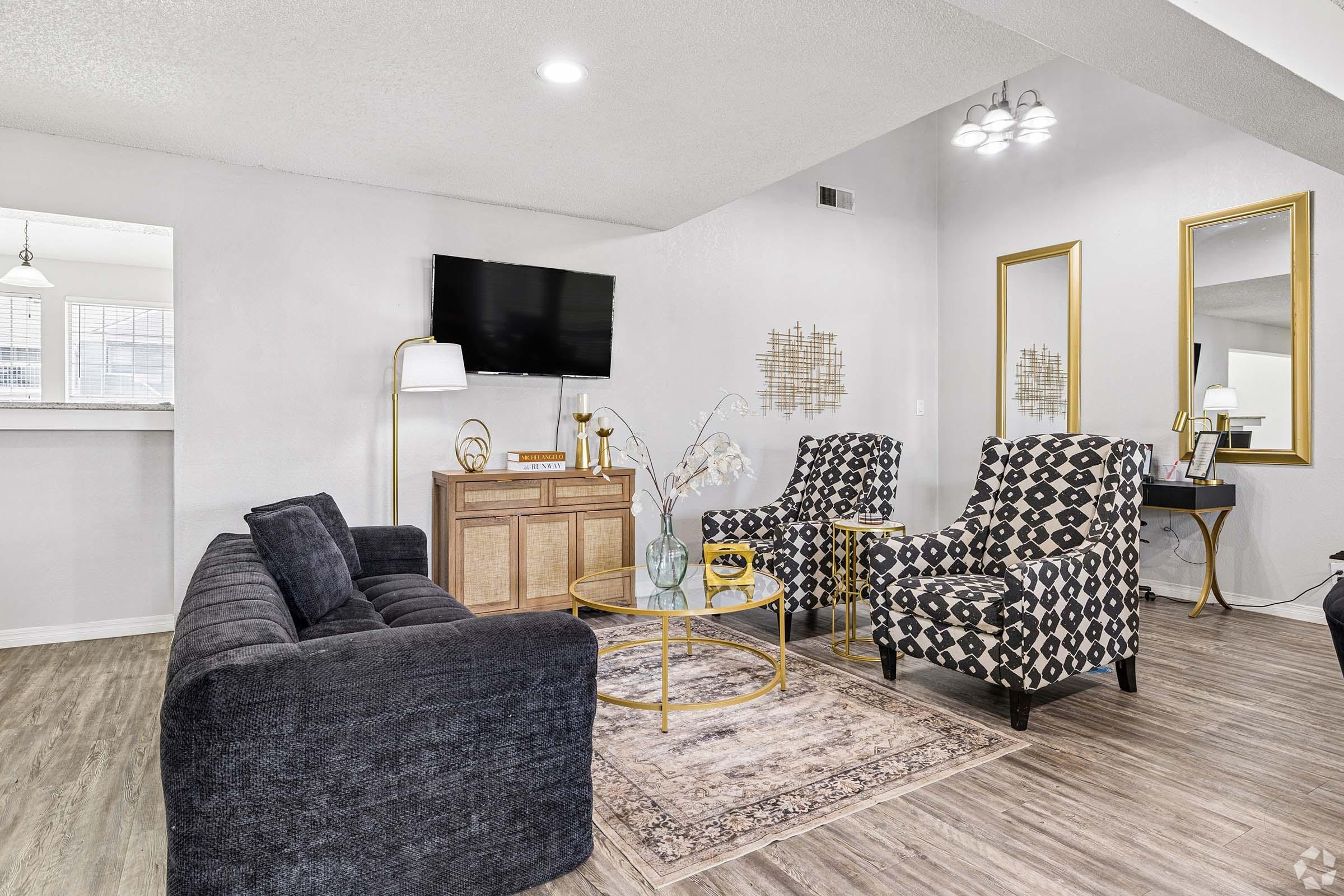 A modern living room featuring a dark gray sofa, two patterned armchairs, and a round coffee table. There's a television mounted on the wall and decorative elements like a gold-framed mirror, a console table, and a rug on a wood-style floor. Natural light filters through the windows.